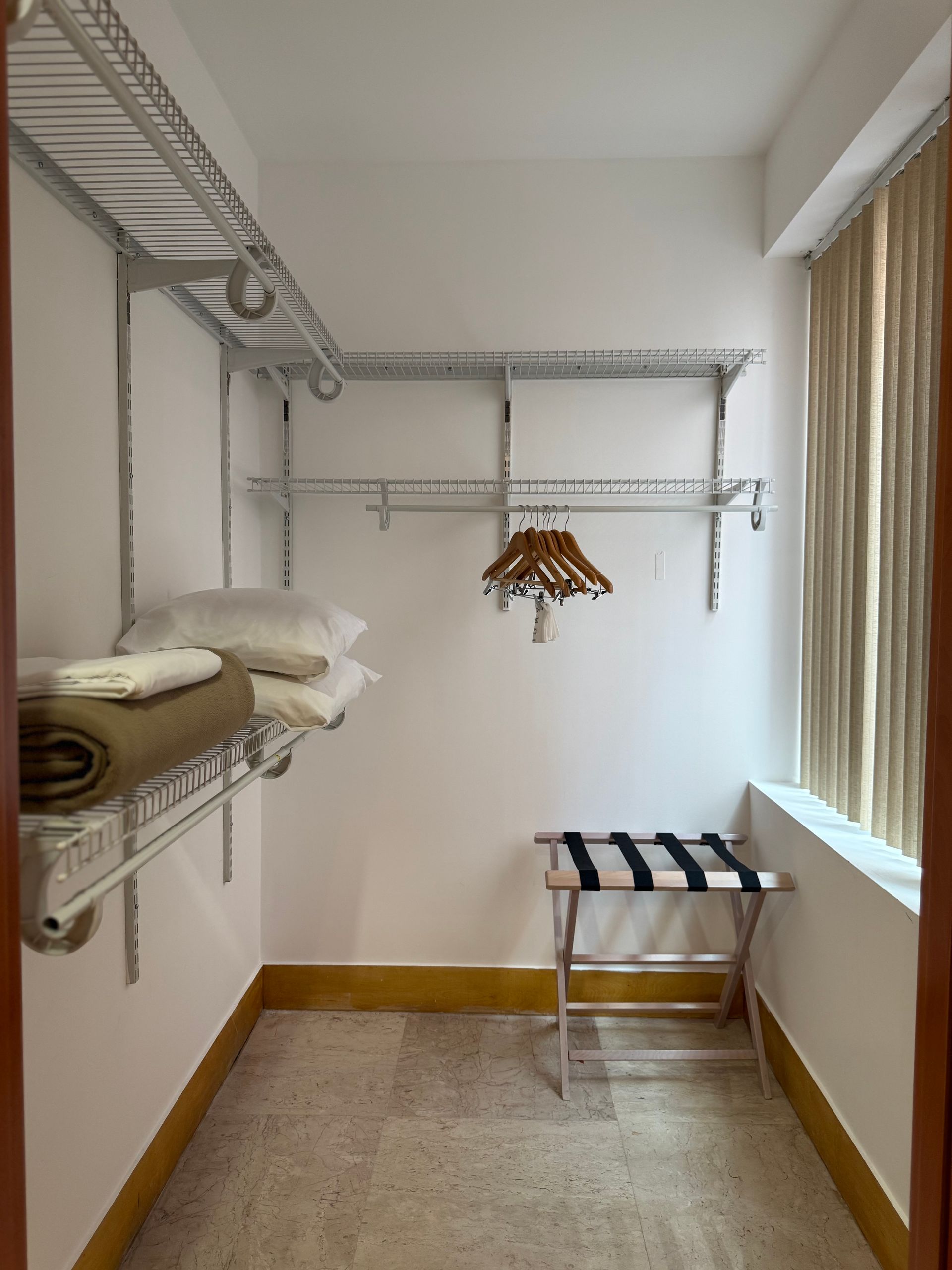 Walk-in closet with wire shelving, folded blankets, pillows, a garment rack, and luggage stand. Beige walls and window.