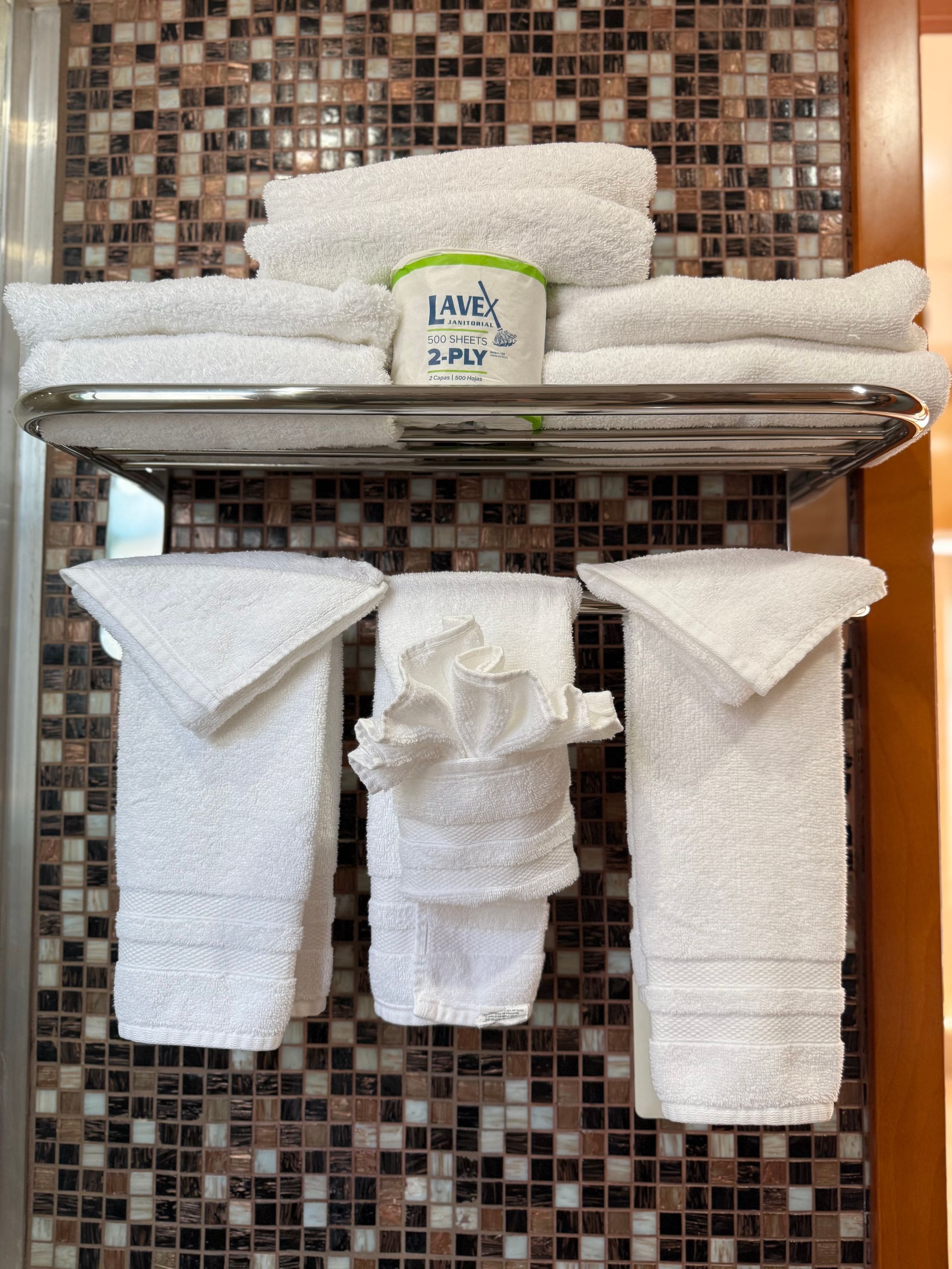 White towels on a chrome rack in a bathroom, with a small container and decorative tiled wall.