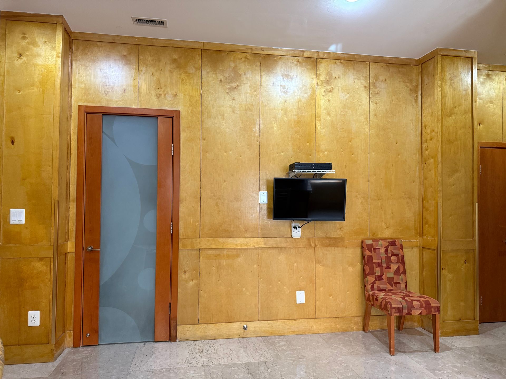 Room with wood paneling, a TV, door, and a patterned chair.