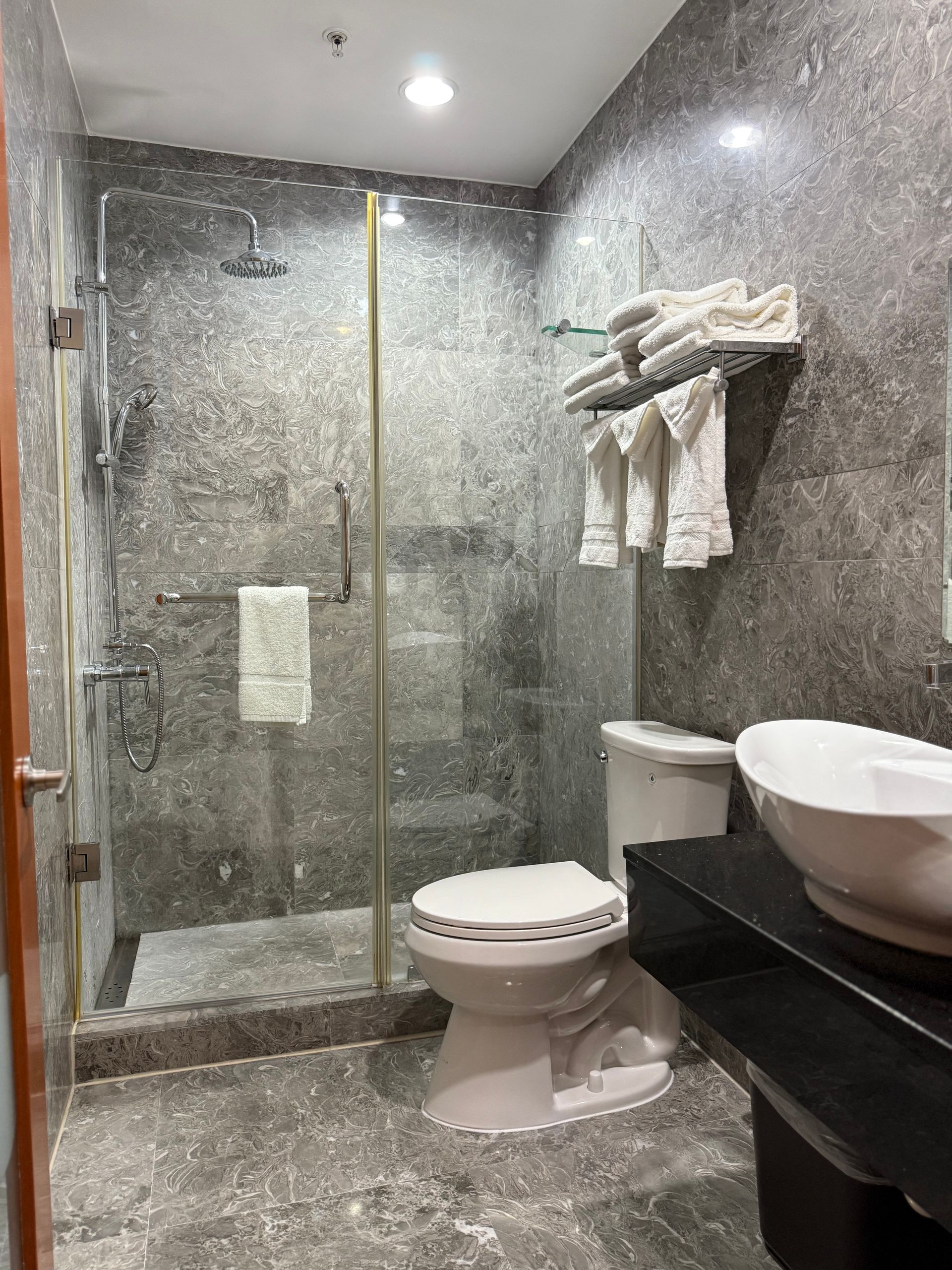 Bathroom with gray speckled walls, glass shower, toilet, white towels, and a vessel sink.