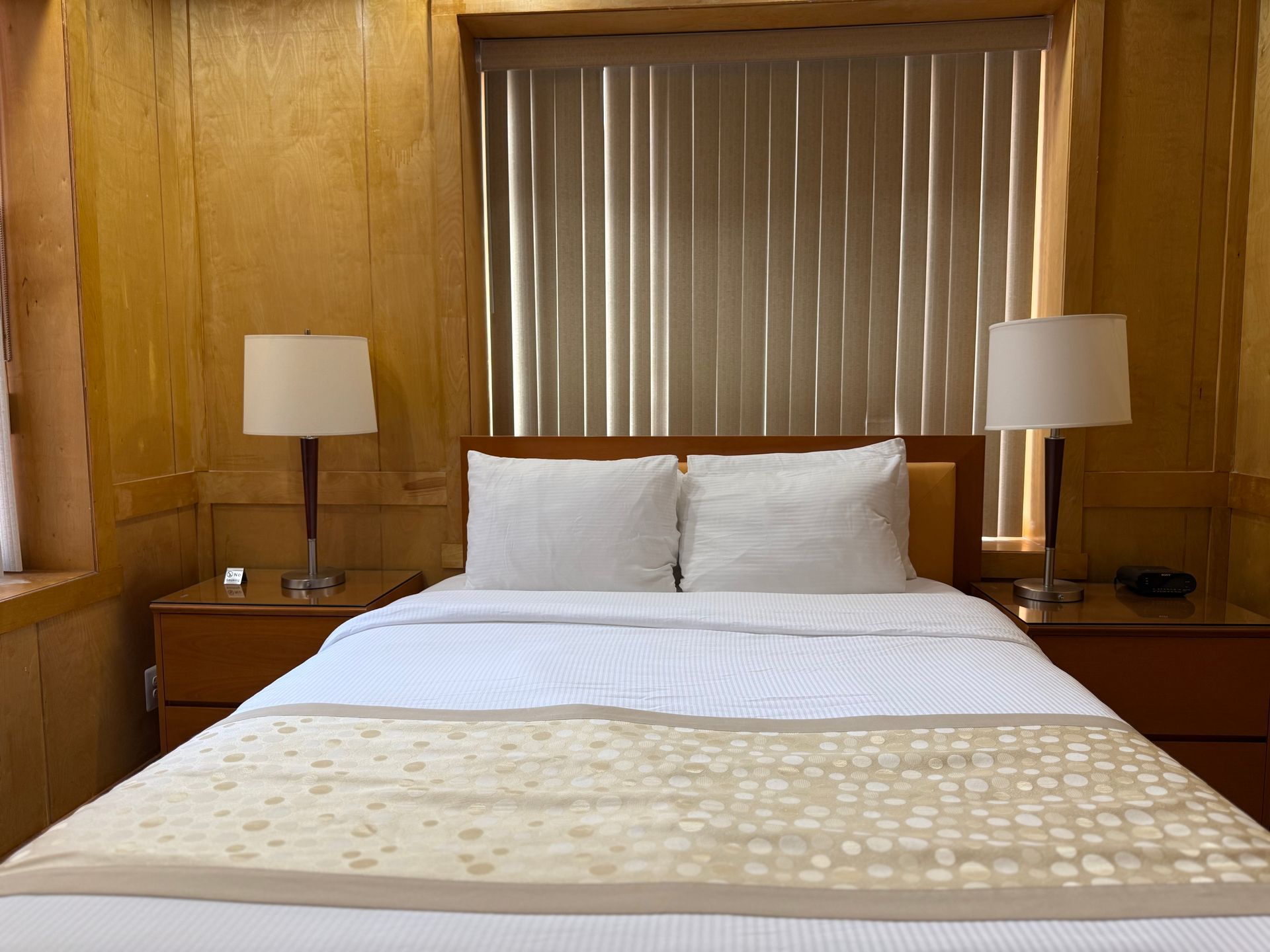 Cozy hotel bedroom with a made bed, two lamps, and window blinds. Warm tones.