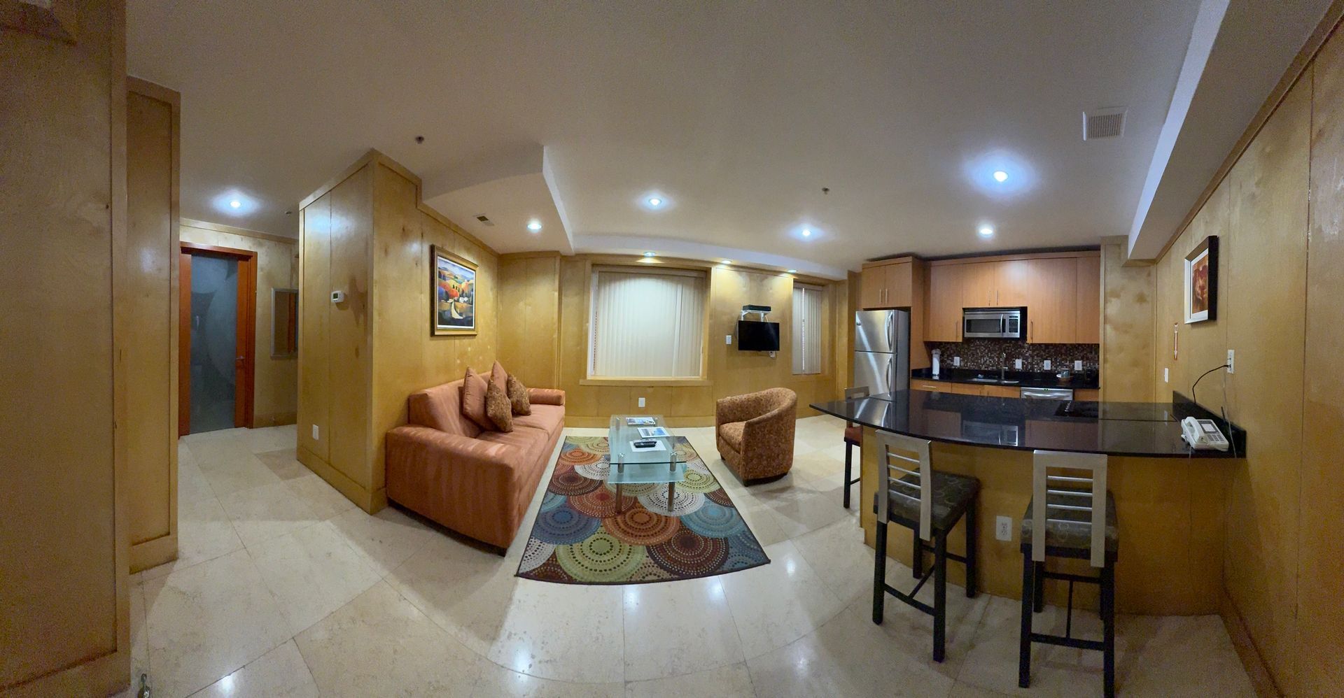 Interior view of a furnished apartment with a kitchen, living area, and bar seating. Wooden paneling throughout.