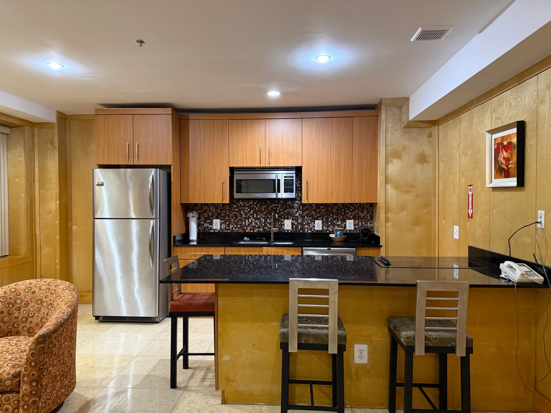 Kitchen with stainless steel refrigerator, wooden cabinets, black granite countertop, and bar stools.