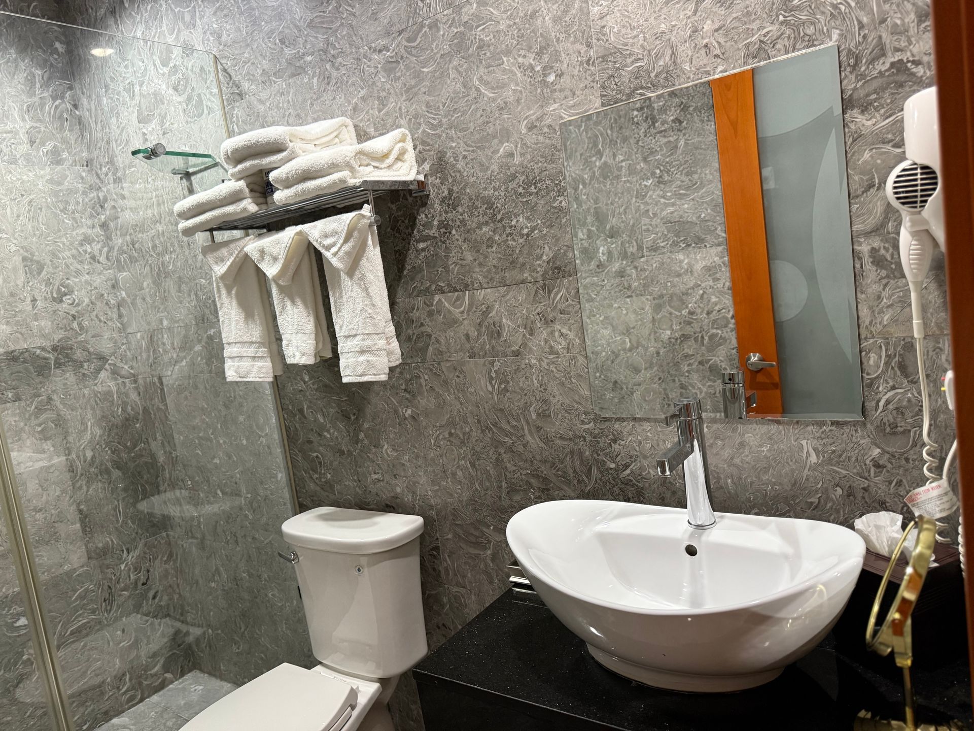 Bathroom with gray tiled walls, white sink, toilet, and towels. Mirror and hairdryer visible.