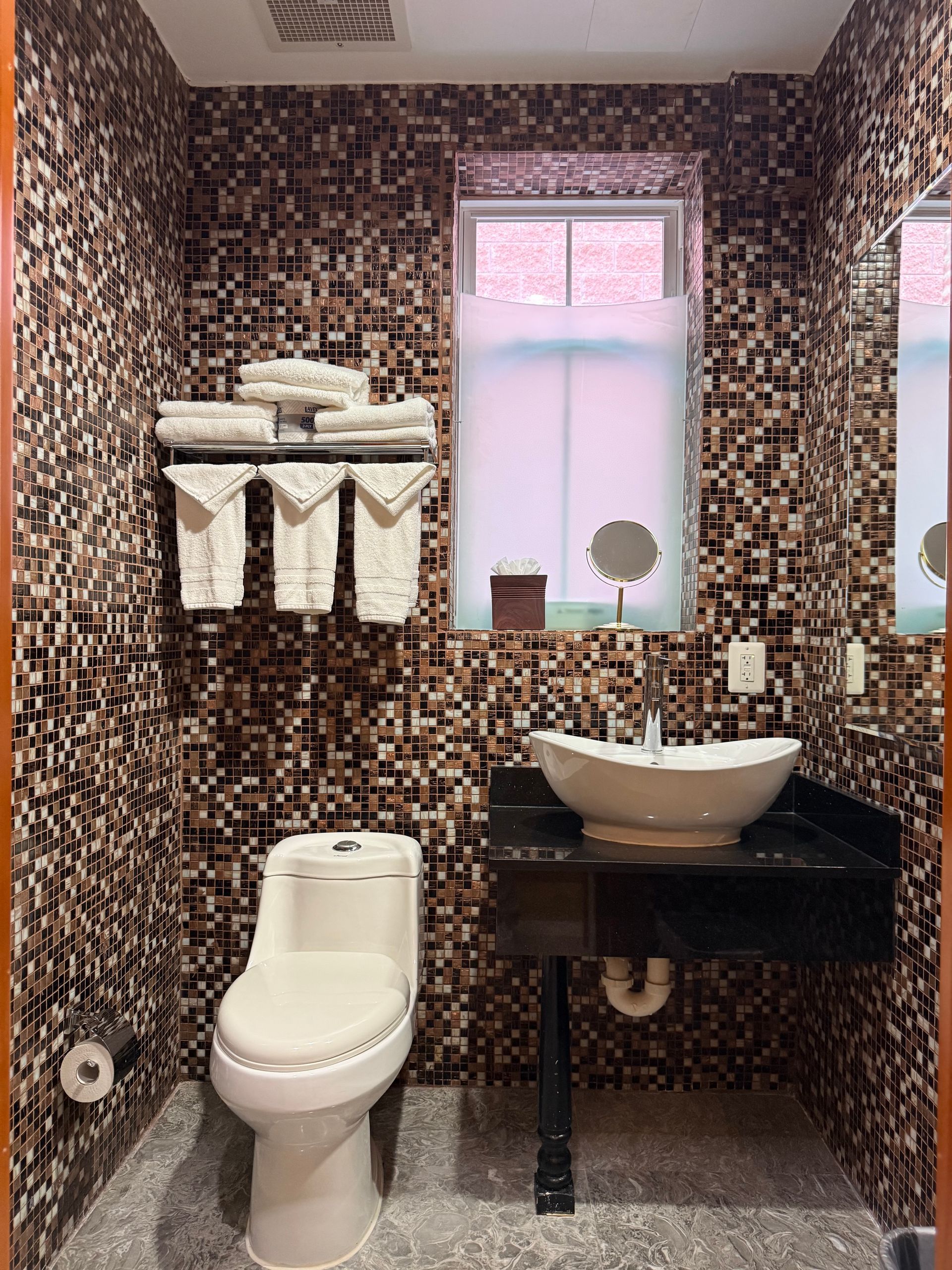Bathroom with mosaic tile walls, a toilet, a vessel sink, and a window with a sheer shade.