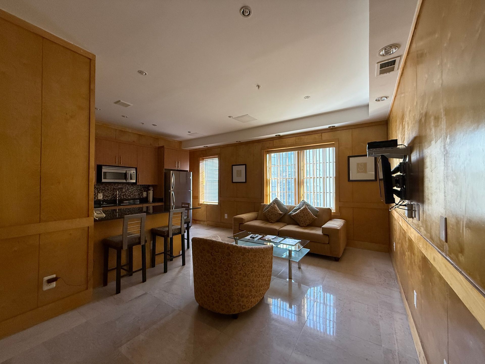 Living room with kitchenette, golden wood paneling, sofa, armchair, TV, bar stools, window.