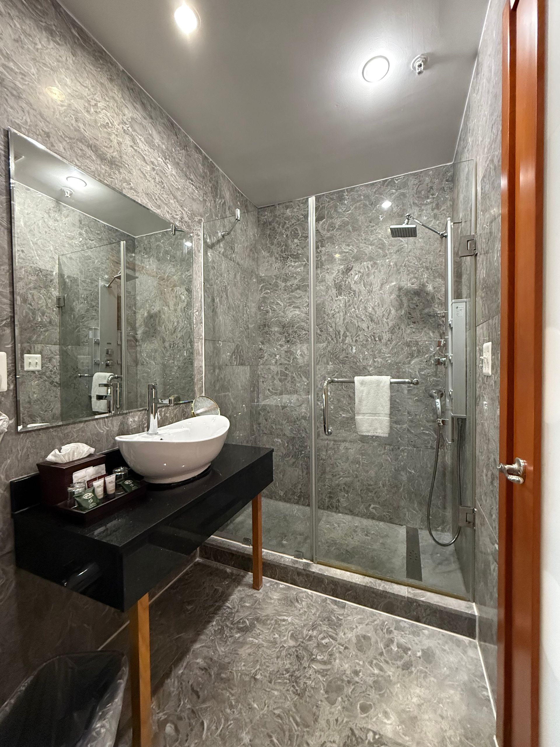 Bathroom with gray stone-like walls and floor, a glass-enclosed shower, a white sink, and a black vanity.