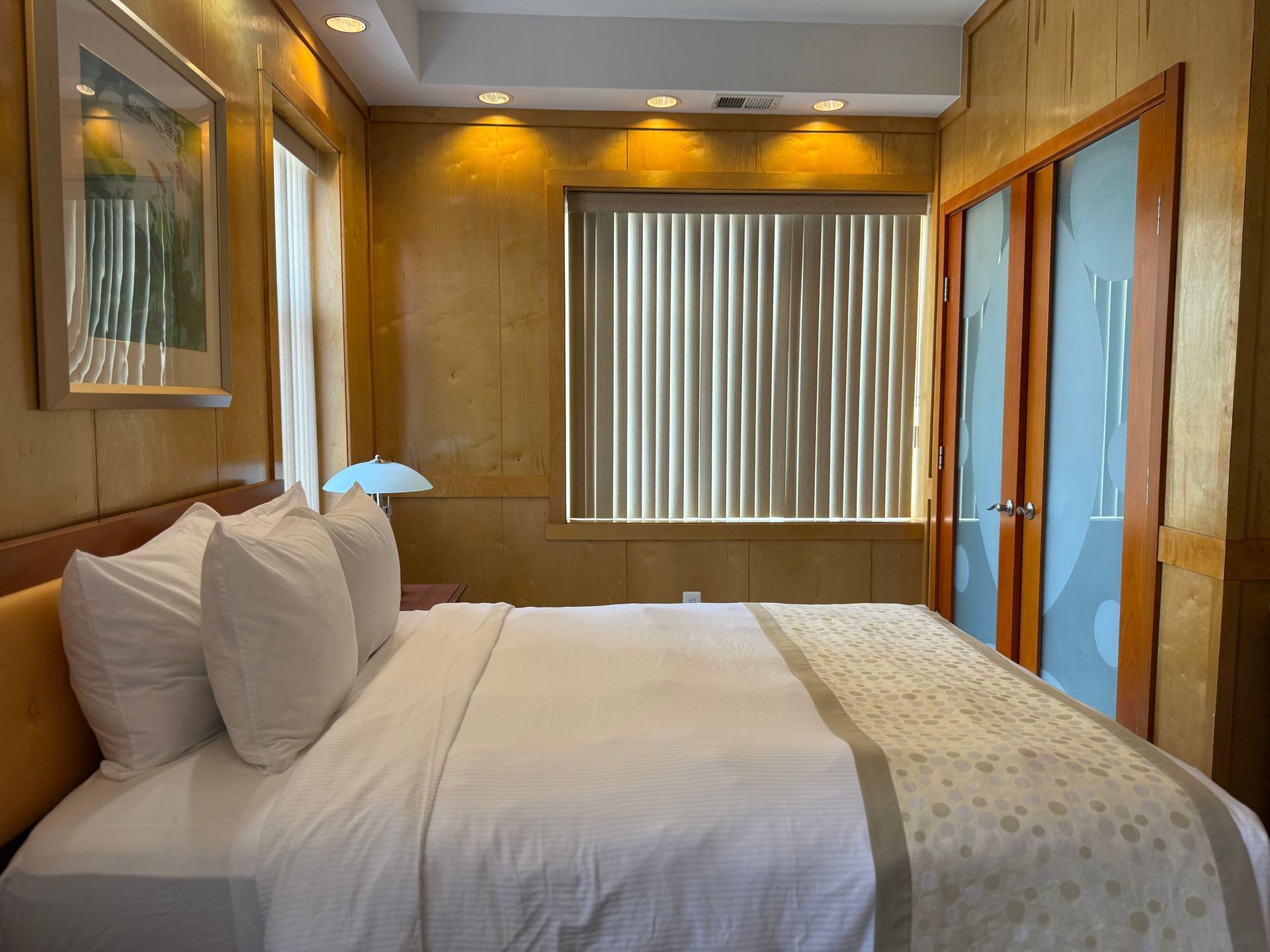 Hotel bedroom with a made bed, window with blinds, and decorative artwork.
