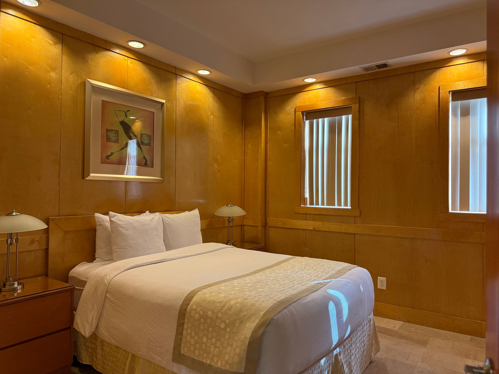 Bedroom with wood-paneled walls, bed, artwork, and small windows with blinds. Soft lighting.