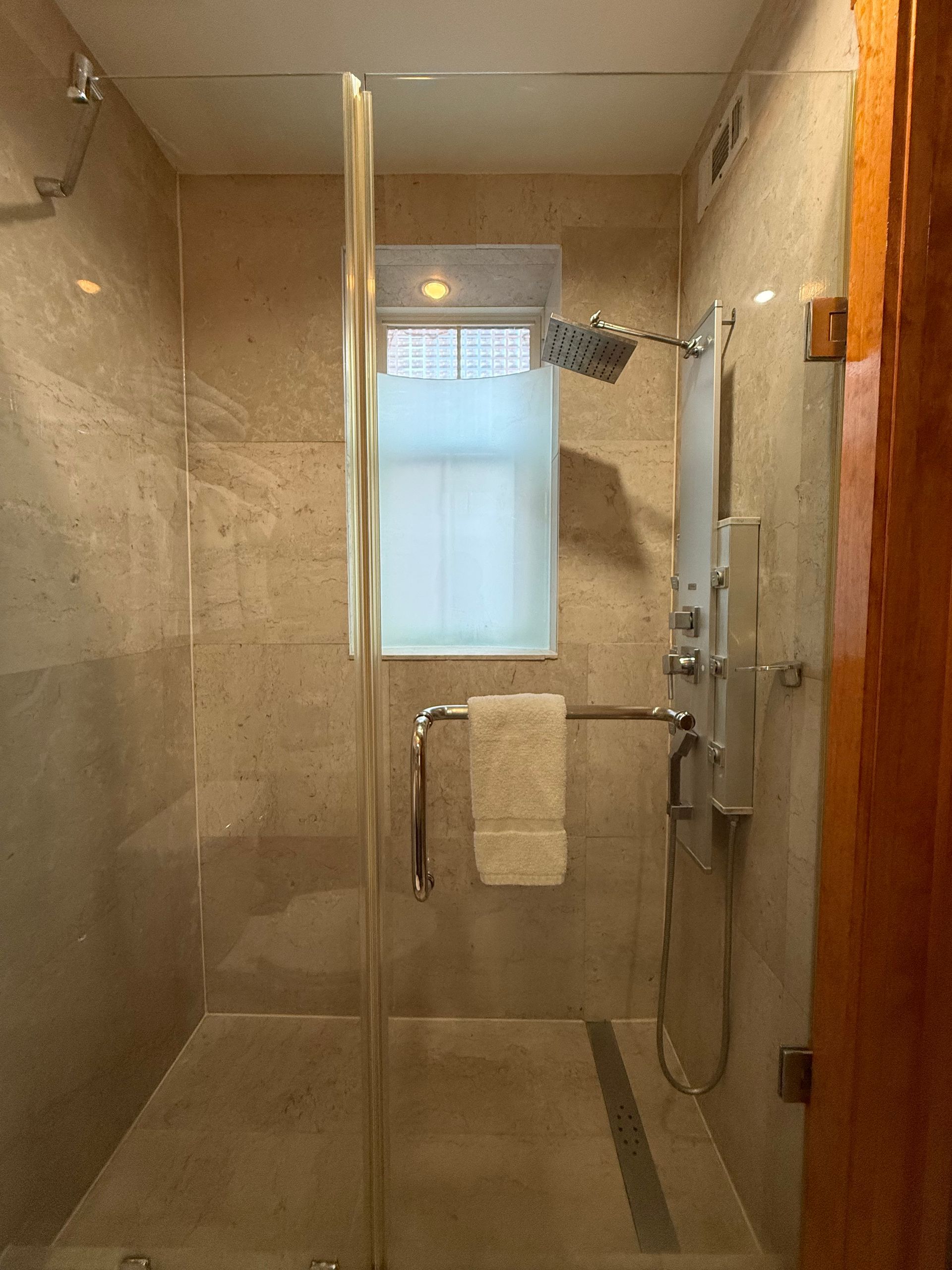 Shower with glass door, small window with frosted glass, showerhead, and a grab bar. Light-colored tiles.