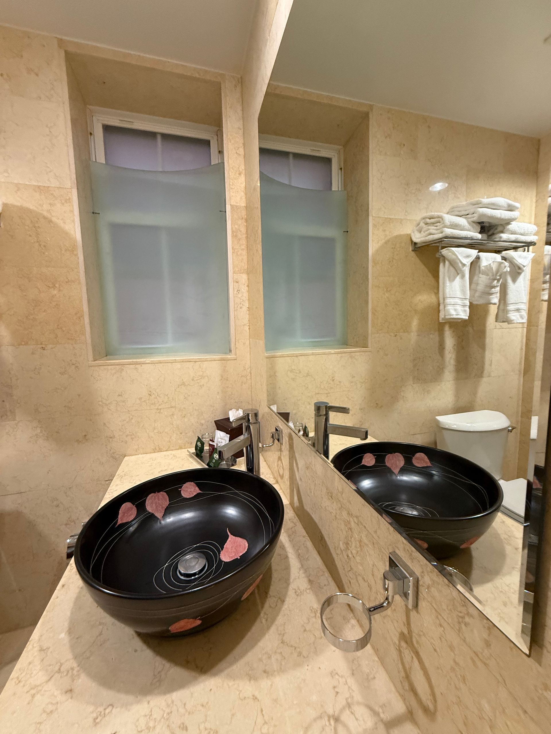 Bathroom with two black bowl sinks, mirror, two frosted windows, beige marble walls.