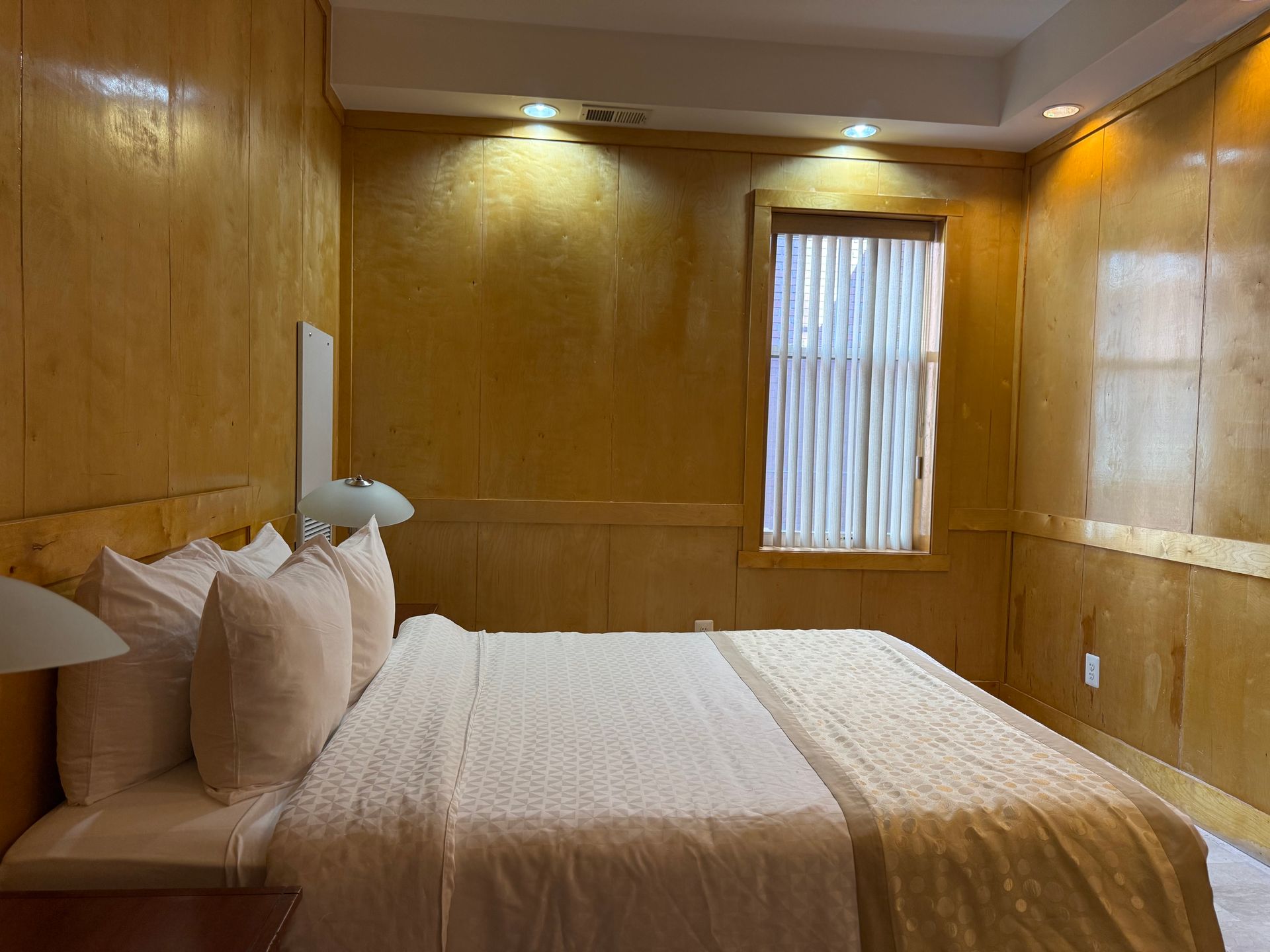 Bedroom with golden wood-paneled walls, bed with white bedding, and a window with blinds.