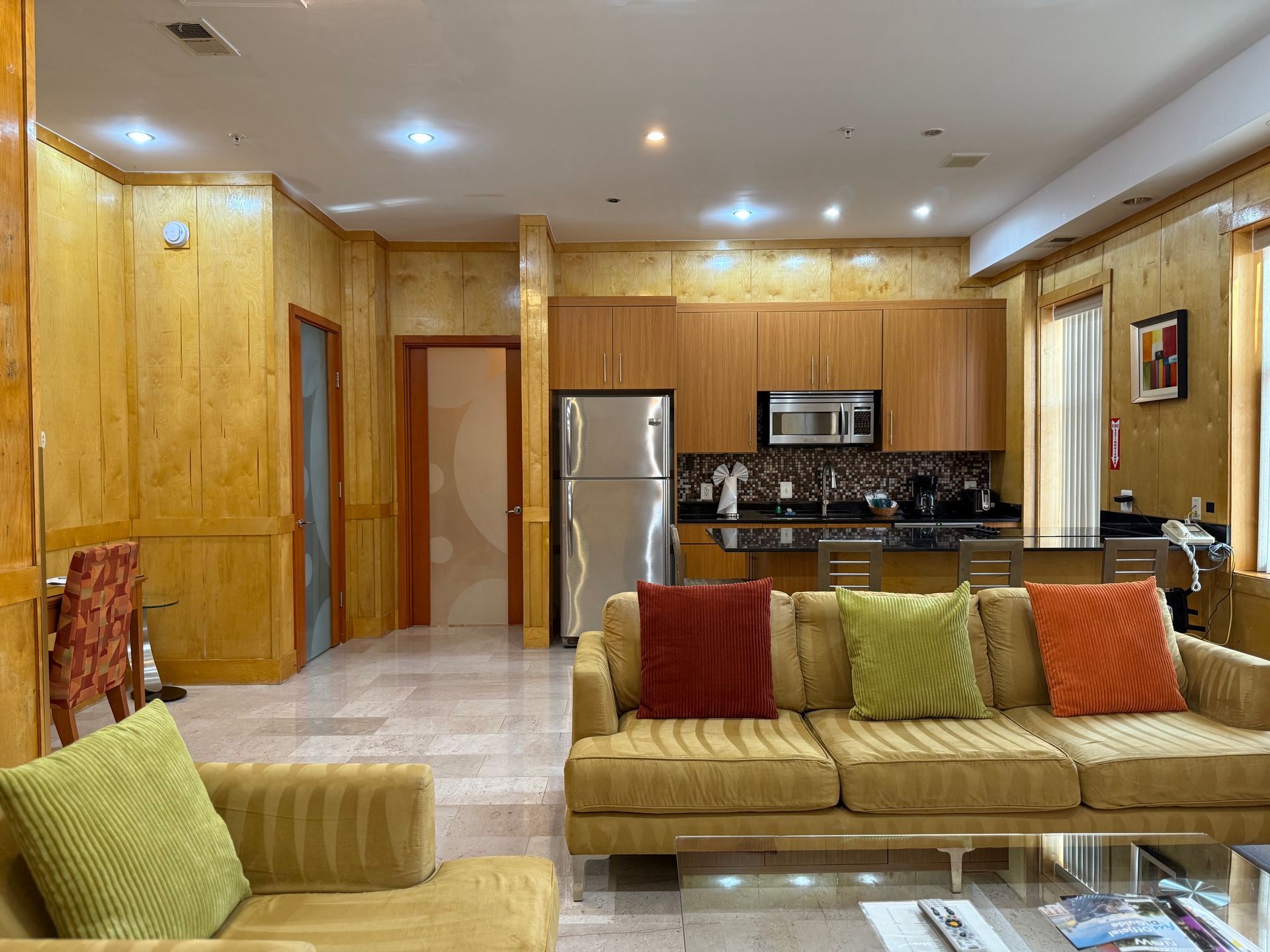 Living area with yellow sofa, kitchen with stainless steel appliances, and wood paneling.