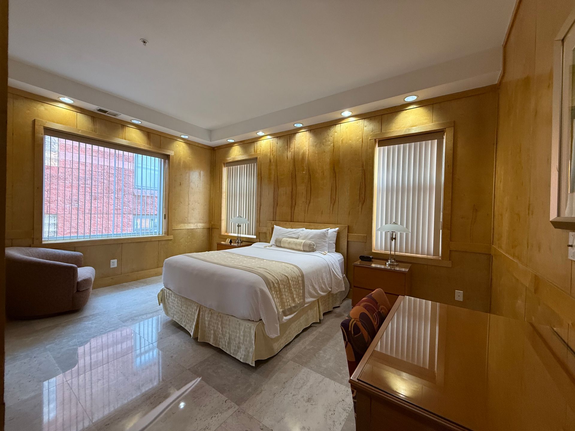 Bedroom with wood-paneled walls, a bed with cream-colored bedding, windows, a chair, and a desk.