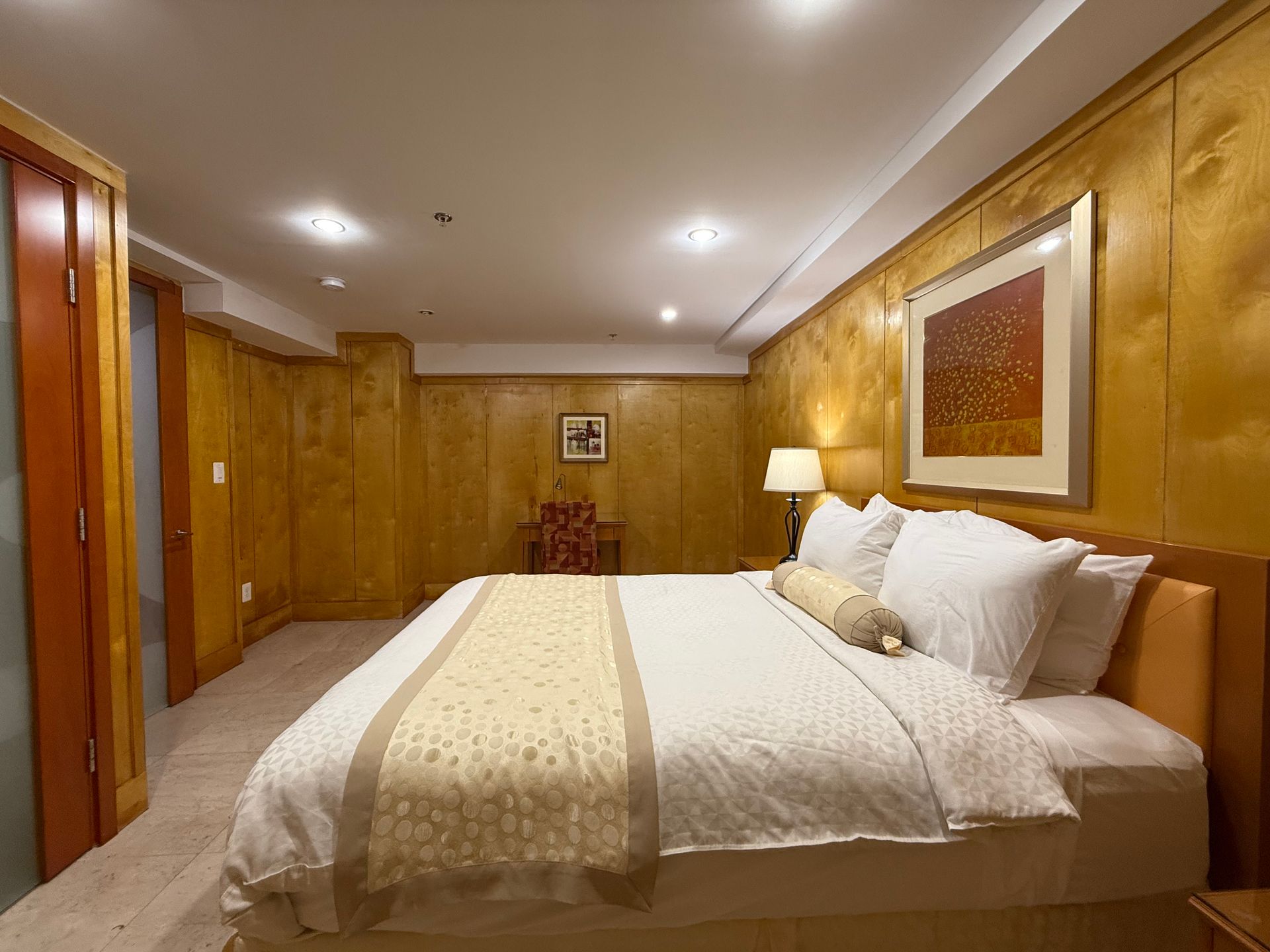 Bedroom with a large bed, gold-toned paneling, a small chair, artwork, and a bedside lamp.