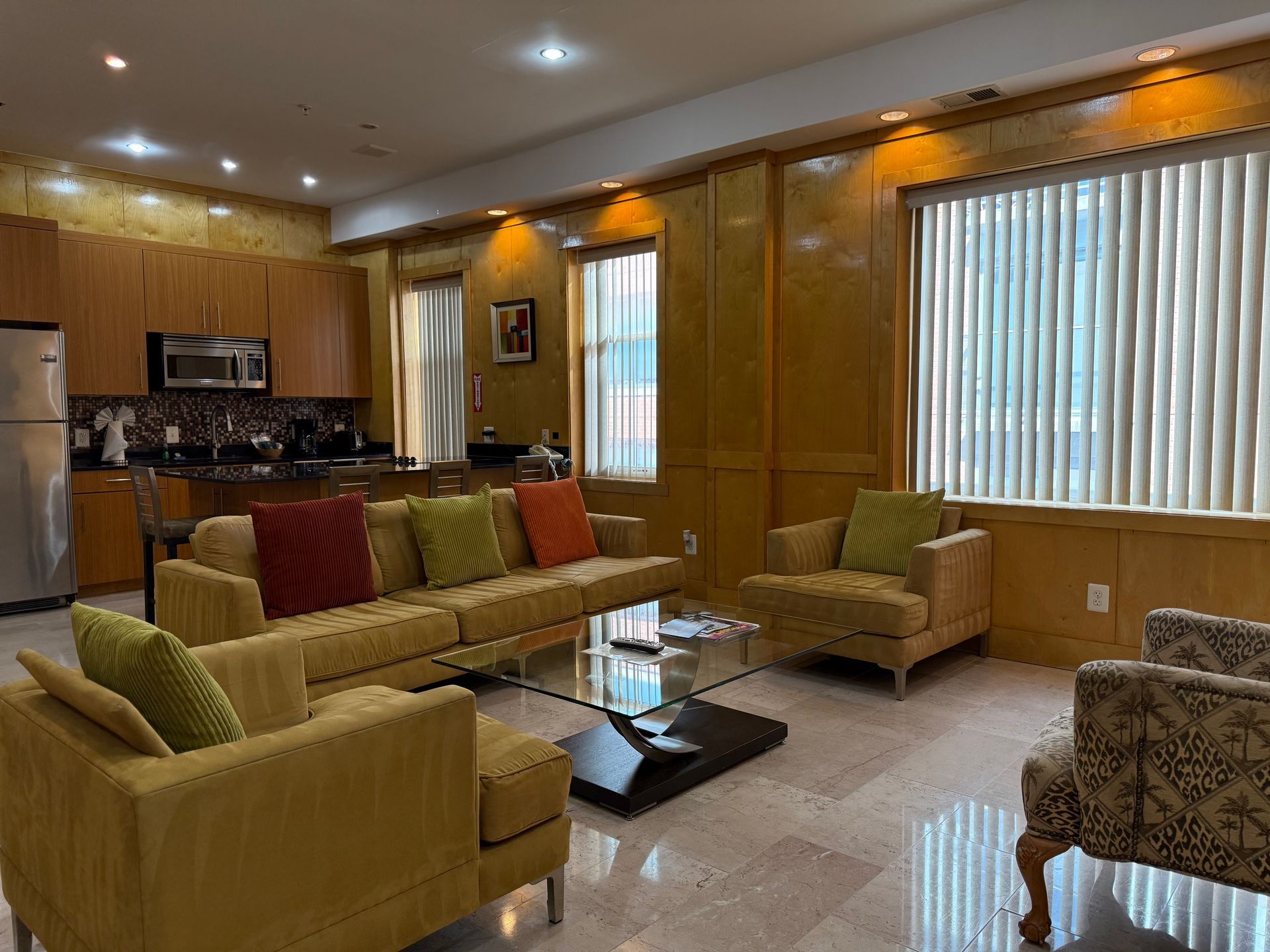 Living room with yellow couch and armchair, open kitchen, windows, and marble floor.