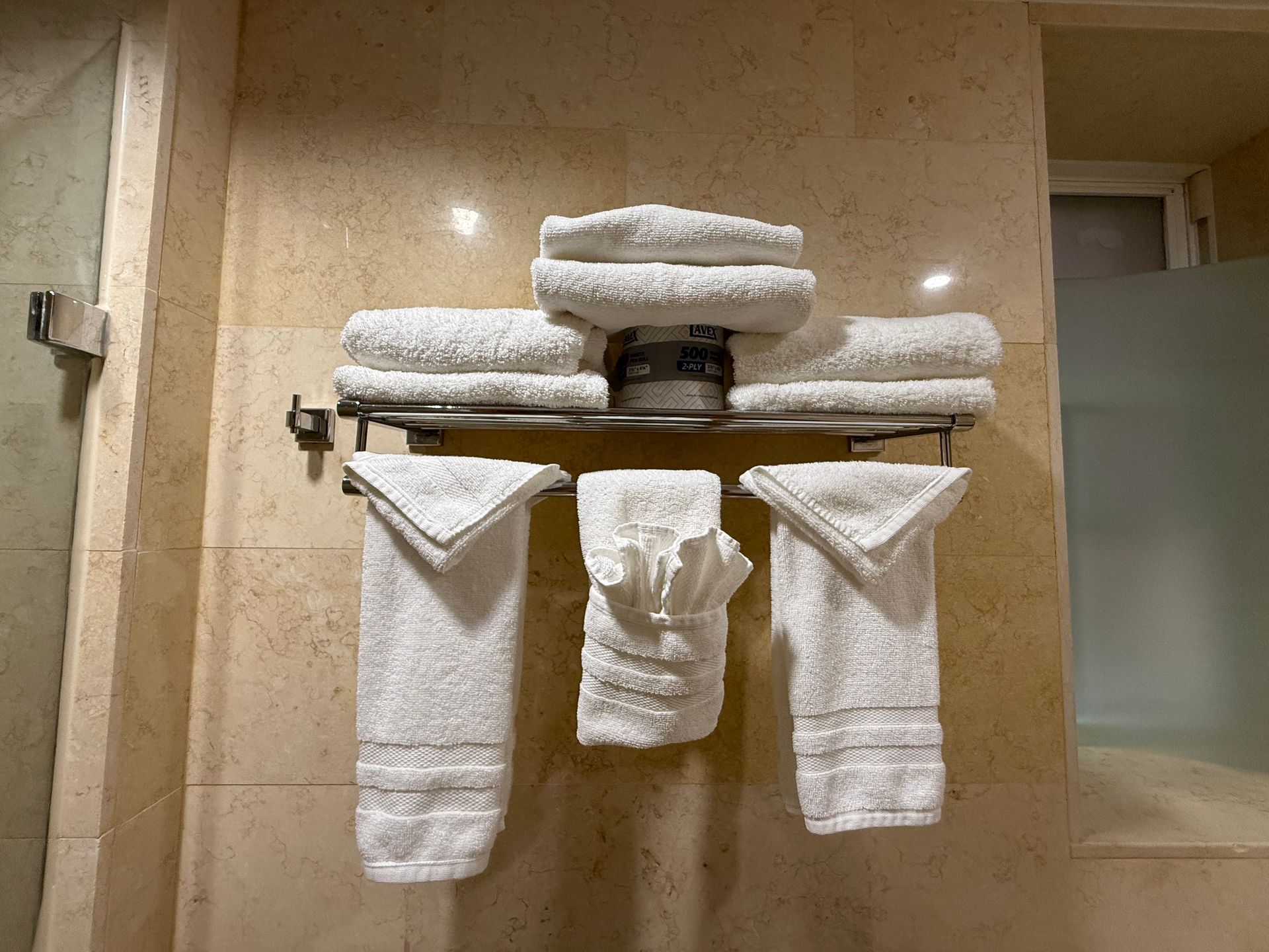 White towels folded and stacked on a chrome towel rack against a beige tiled wall.