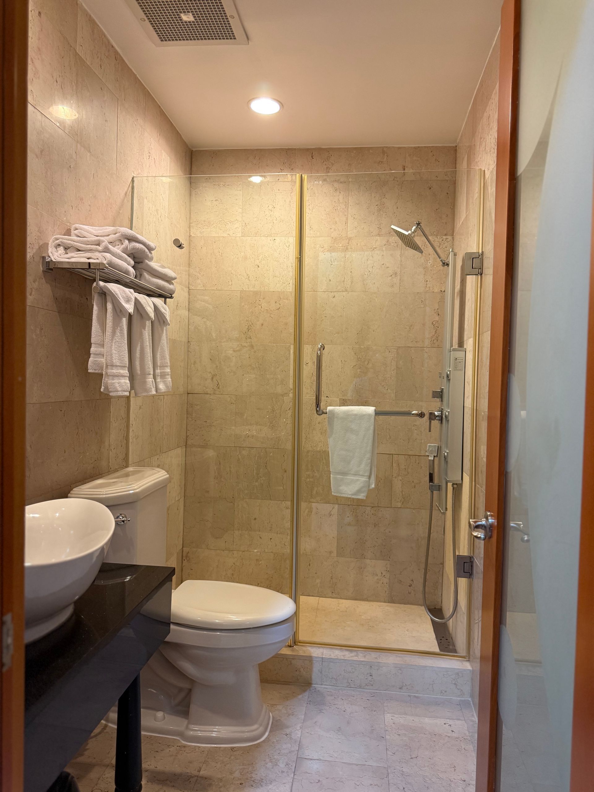 Bathroom with glass shower, toilet, sink, and towels. Light-colored walls and floor.
