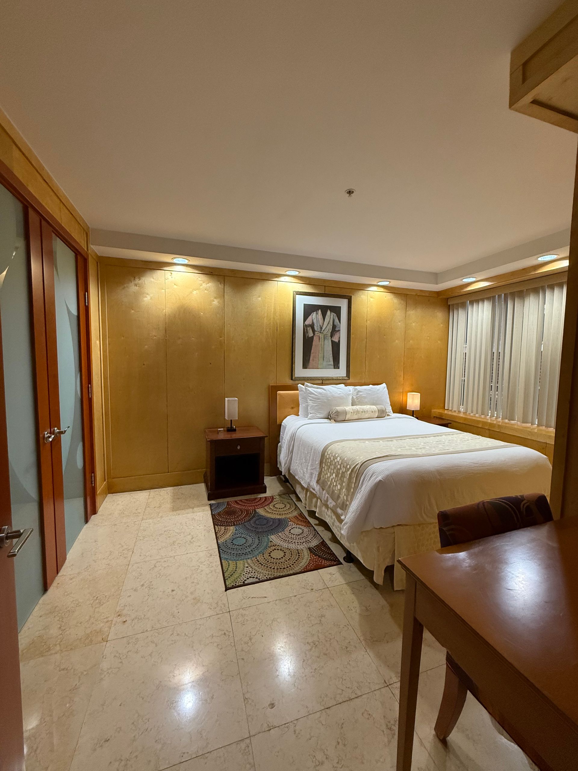 Bedroom with a bed, nightstand, artwork, and a small rug on a light-colored floor. Walls are gold-toned.