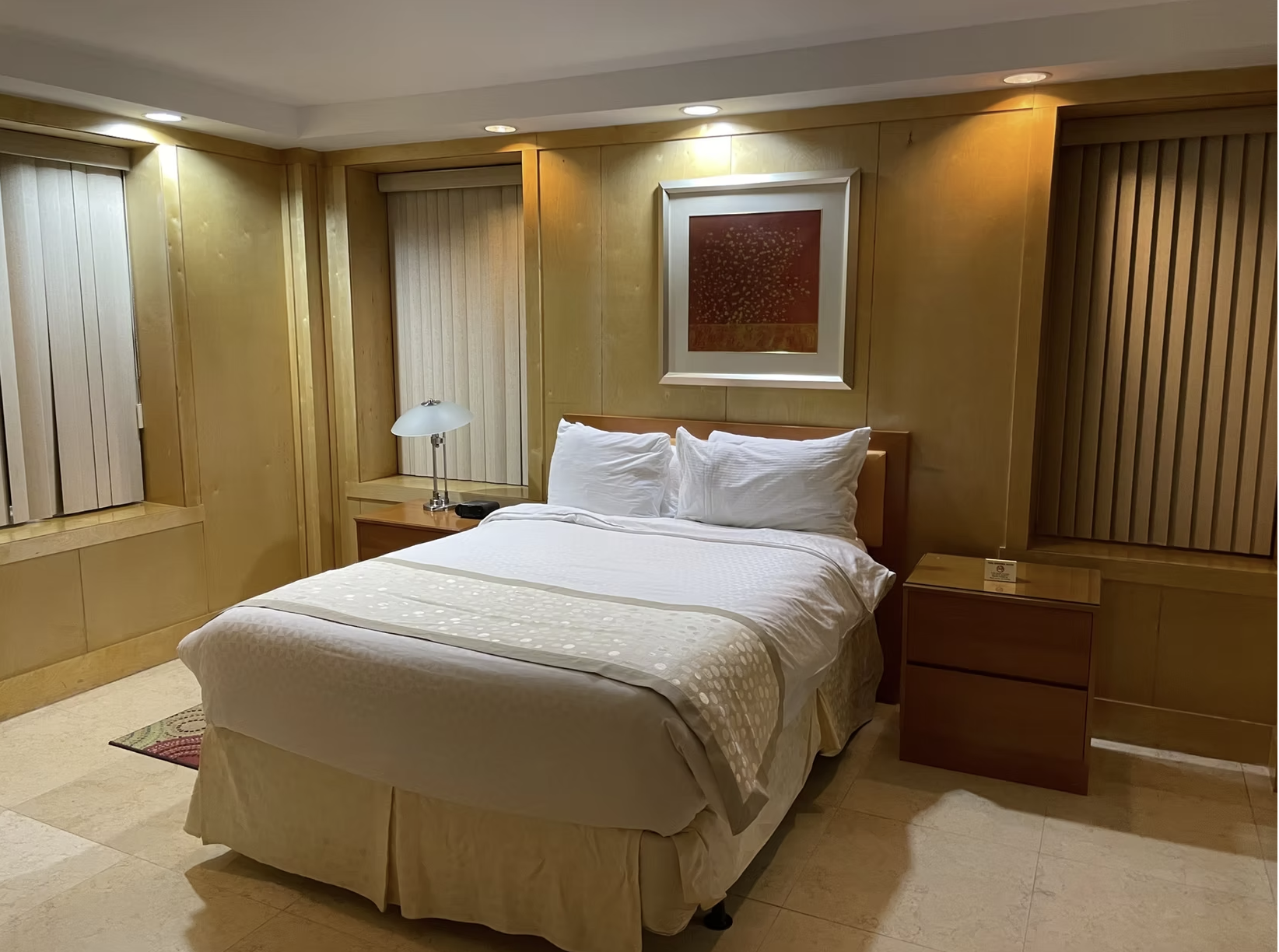 Bedroom with a bed, nightstands, windows with blinds, and artwork; warm lighting.