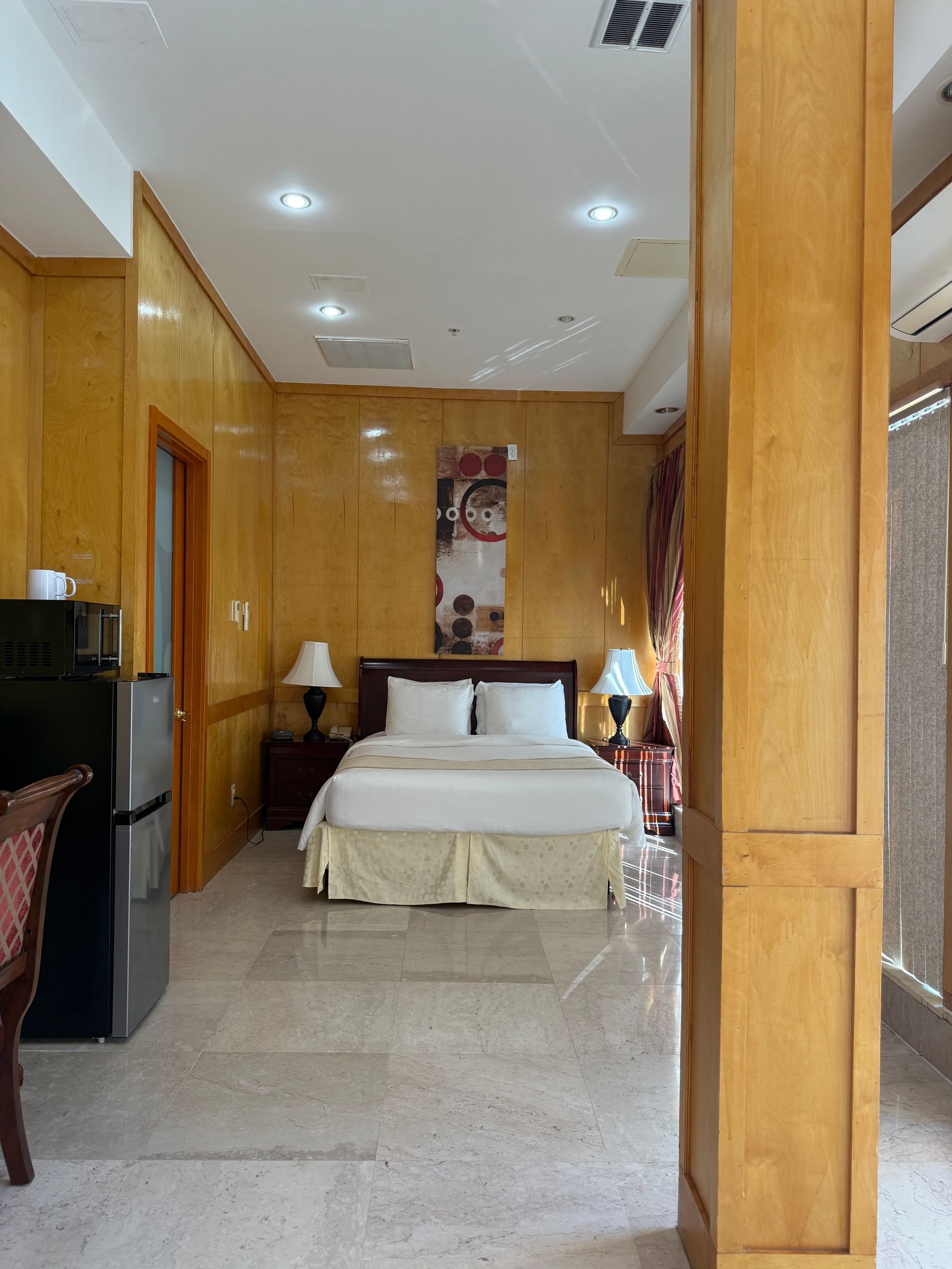 Bedroom with a bed, nightstands, lamps, and a small refrigerator. Shiny, light-colored wood paneling on the walls.