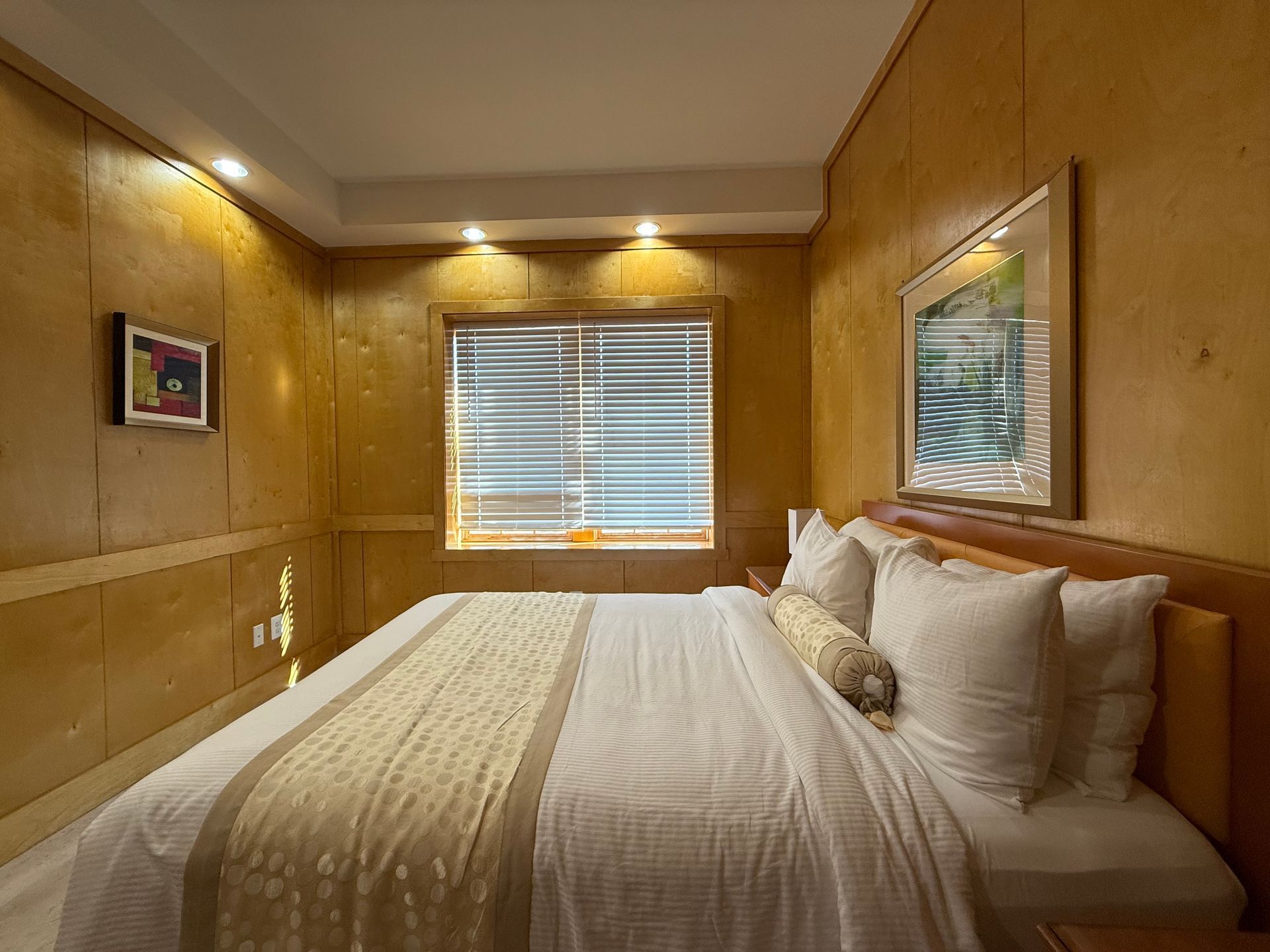 Bedroom with a large bed, window with blinds, artwork, and wood paneling.