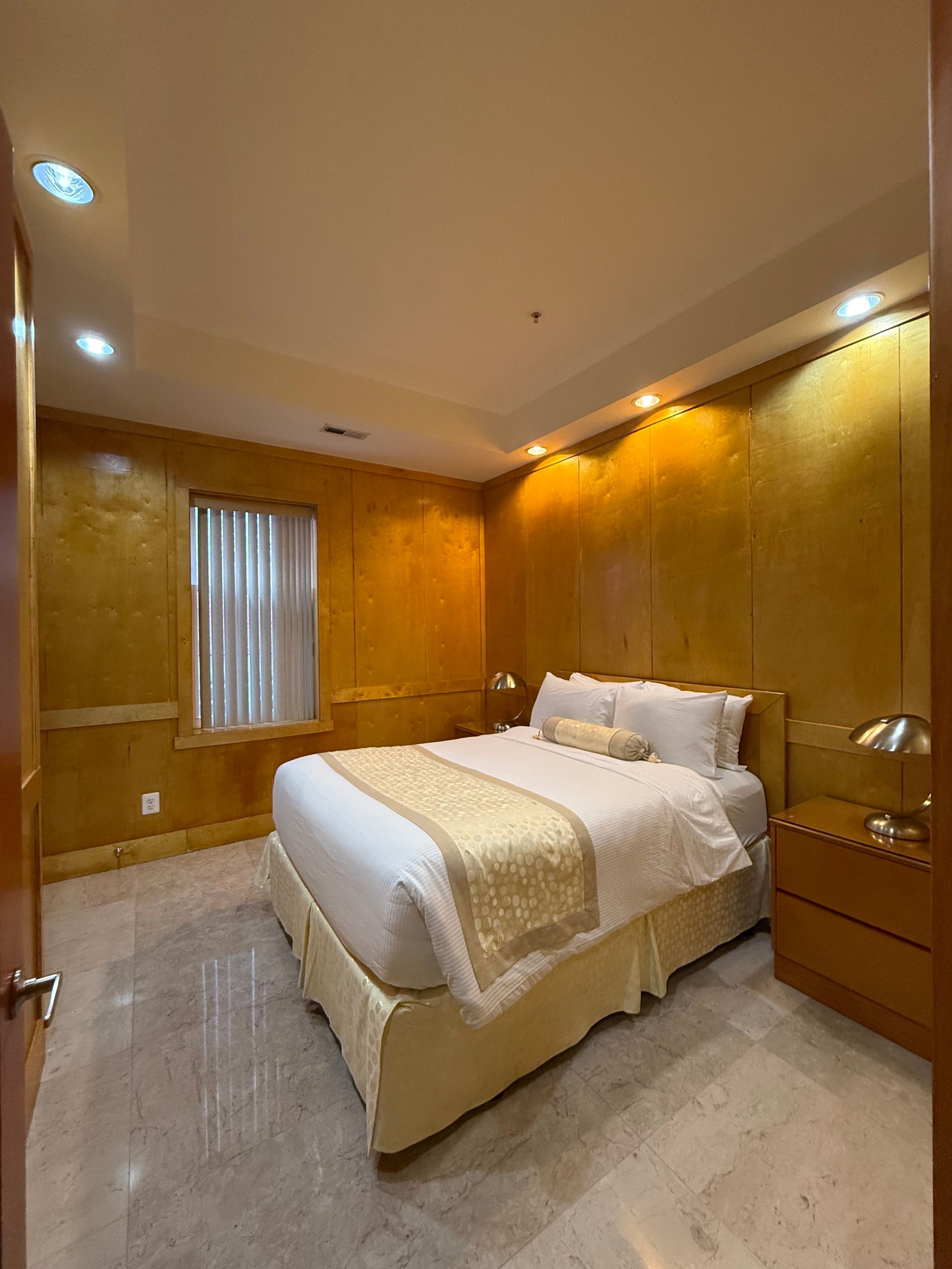 Bedroom with gold-toned walls, cream bedding, and marble floor. A bed is centered, flanked by nightstands.
