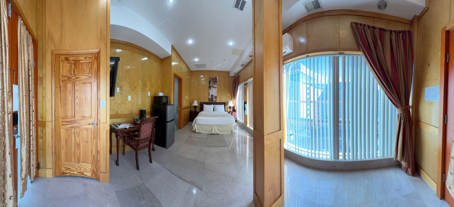 Hotel room interior with bed, desk, and large window. Wooden paneling and tile floor.