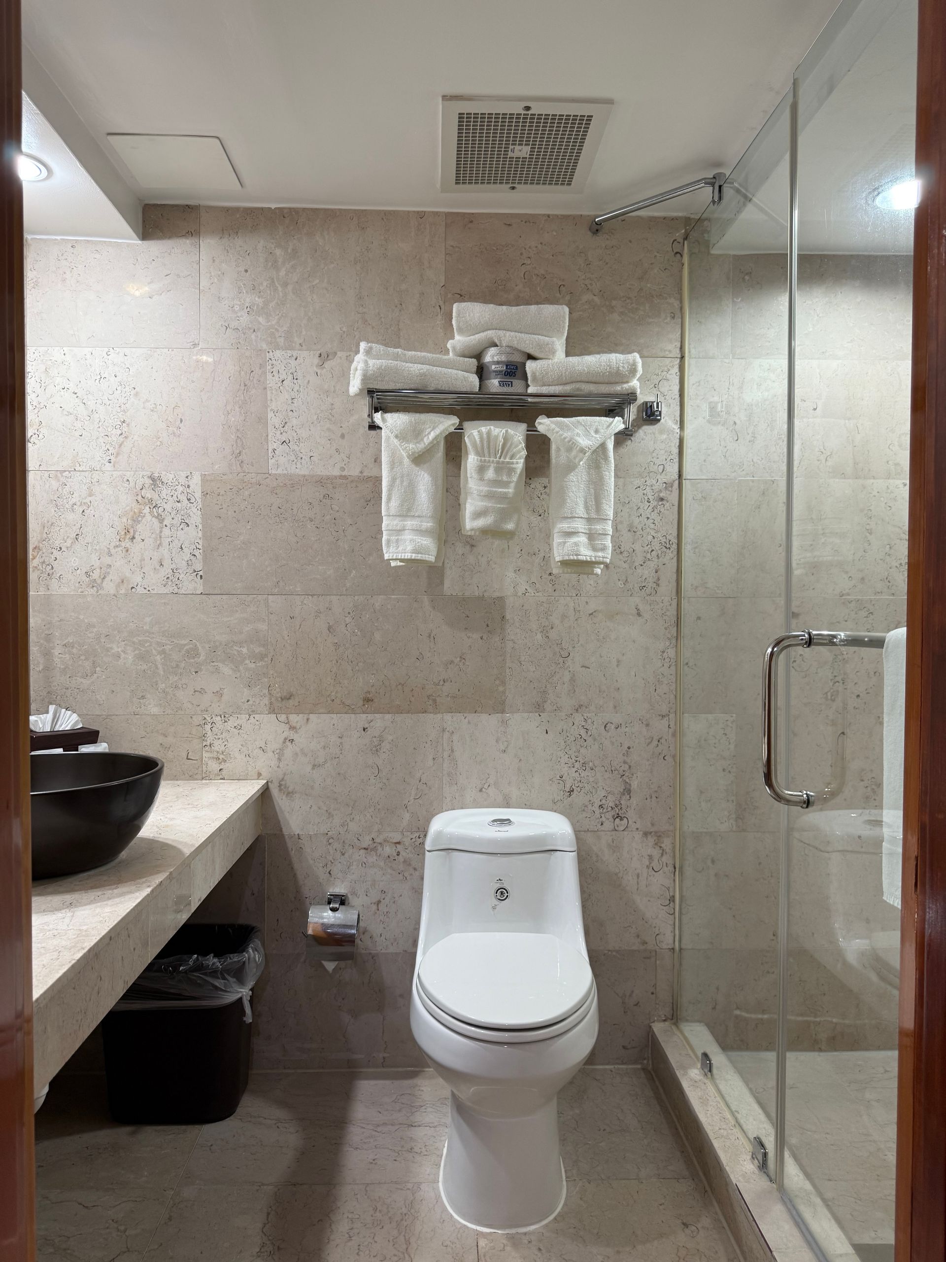Bathroom with a toilet, shower, sink, and towels on a rack. Stone-like tile walls and a glass shower door.