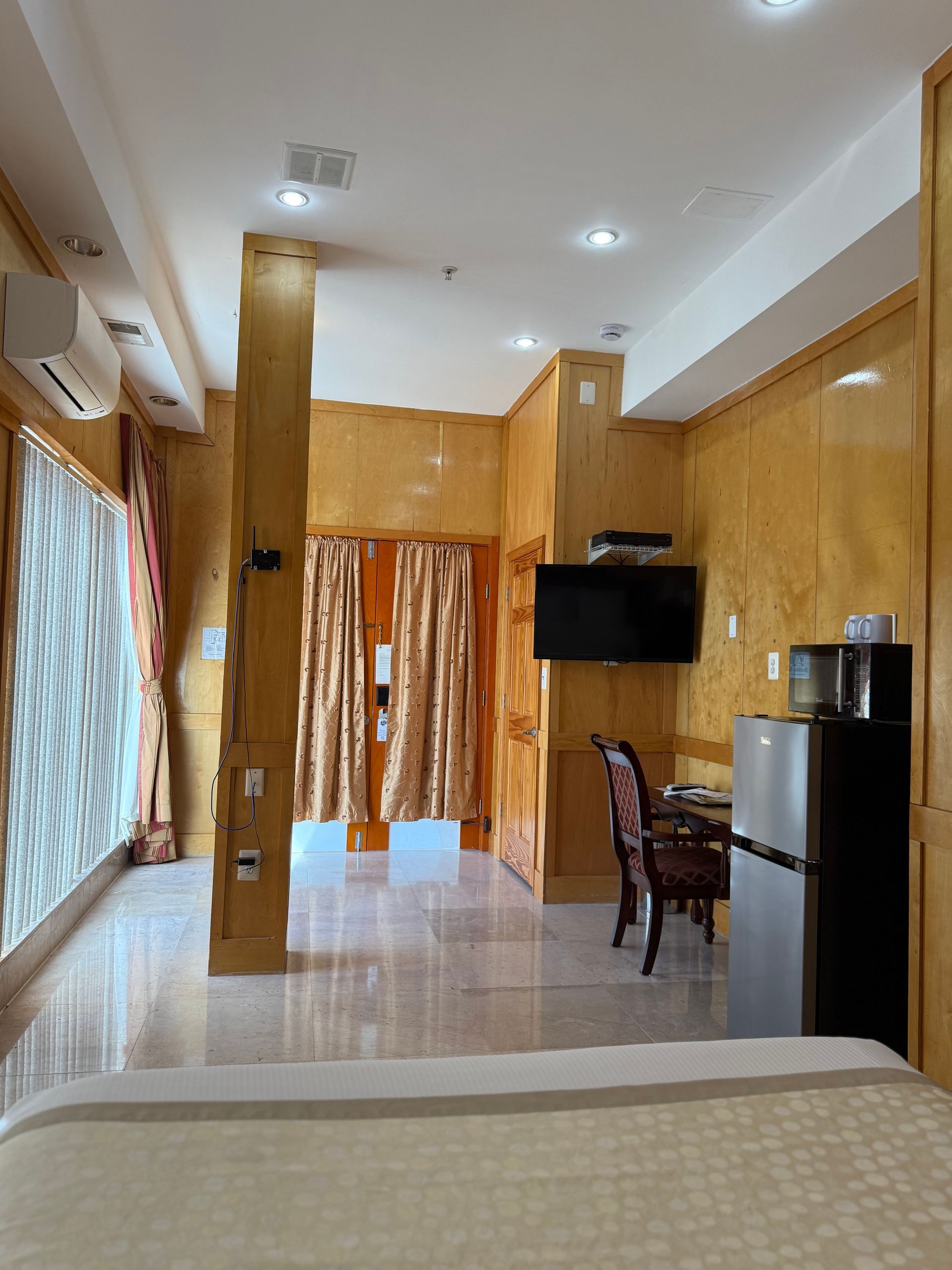 Small hotel room with a TV, desk, refrigerator, and doorway. Walls are gold-colored.