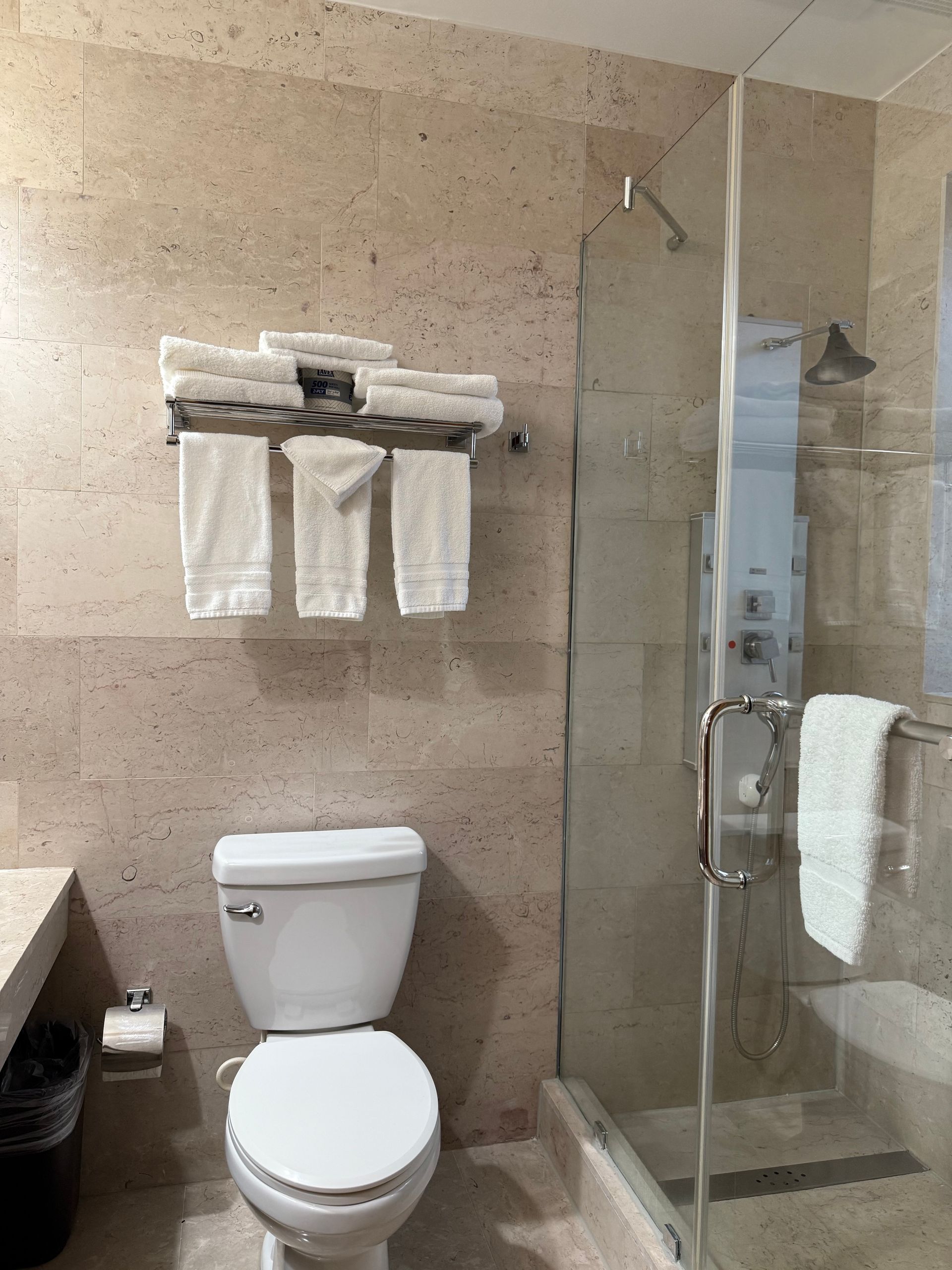 Bathroom with toilet, shower, and towel rack. Tan tile walls and glass shower door. White towels.