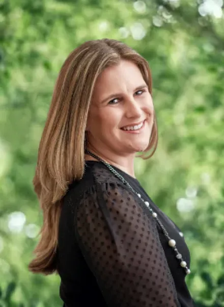 A professional headshot of a smiling woman with long brown hair, wearing a black polka-dot top, set against a green background.