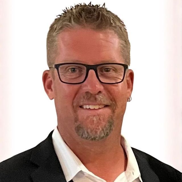 A person with short blond hair and glasses smiles, wearing a white shirt and dark blazer against a plain white background.