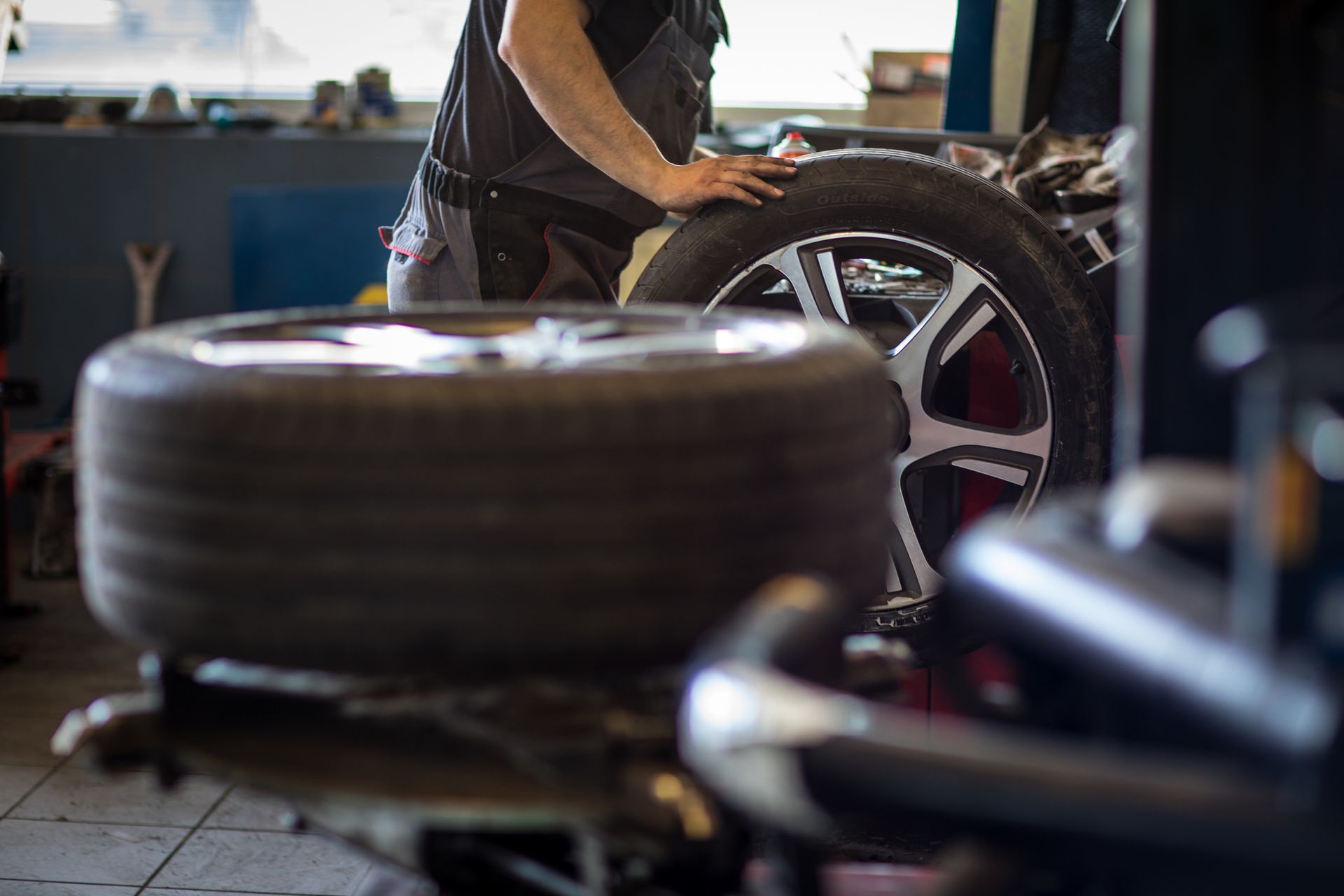 Mechanic in a garage with tires, one mounted on a machine, another in the foreground.