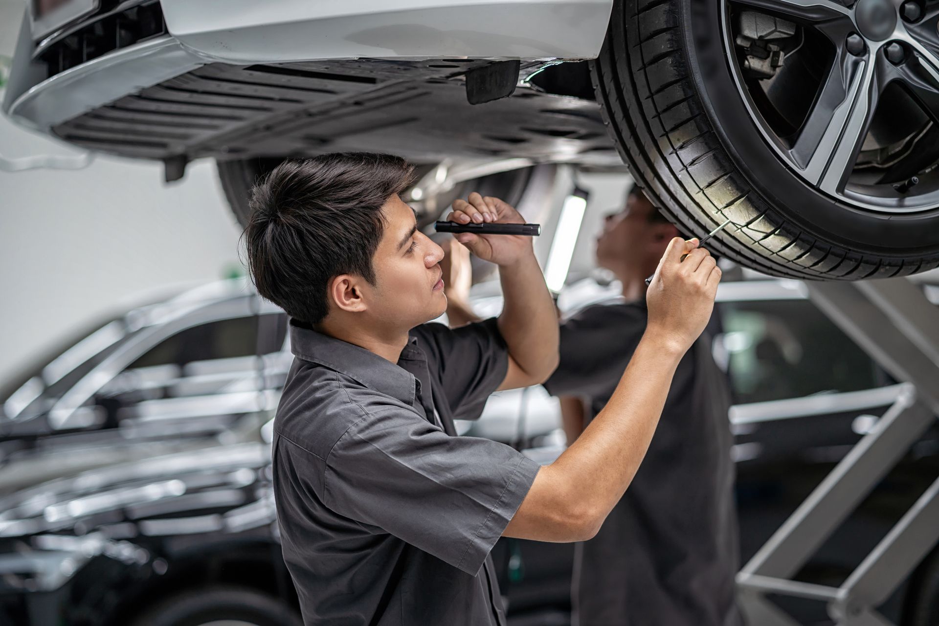 Two mechanics inspecting a car raised on a lift in a garage, using flashlights.