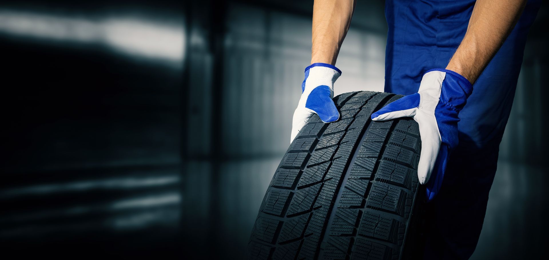 Person in blue uniform and gloves carrying a tire in a garage.