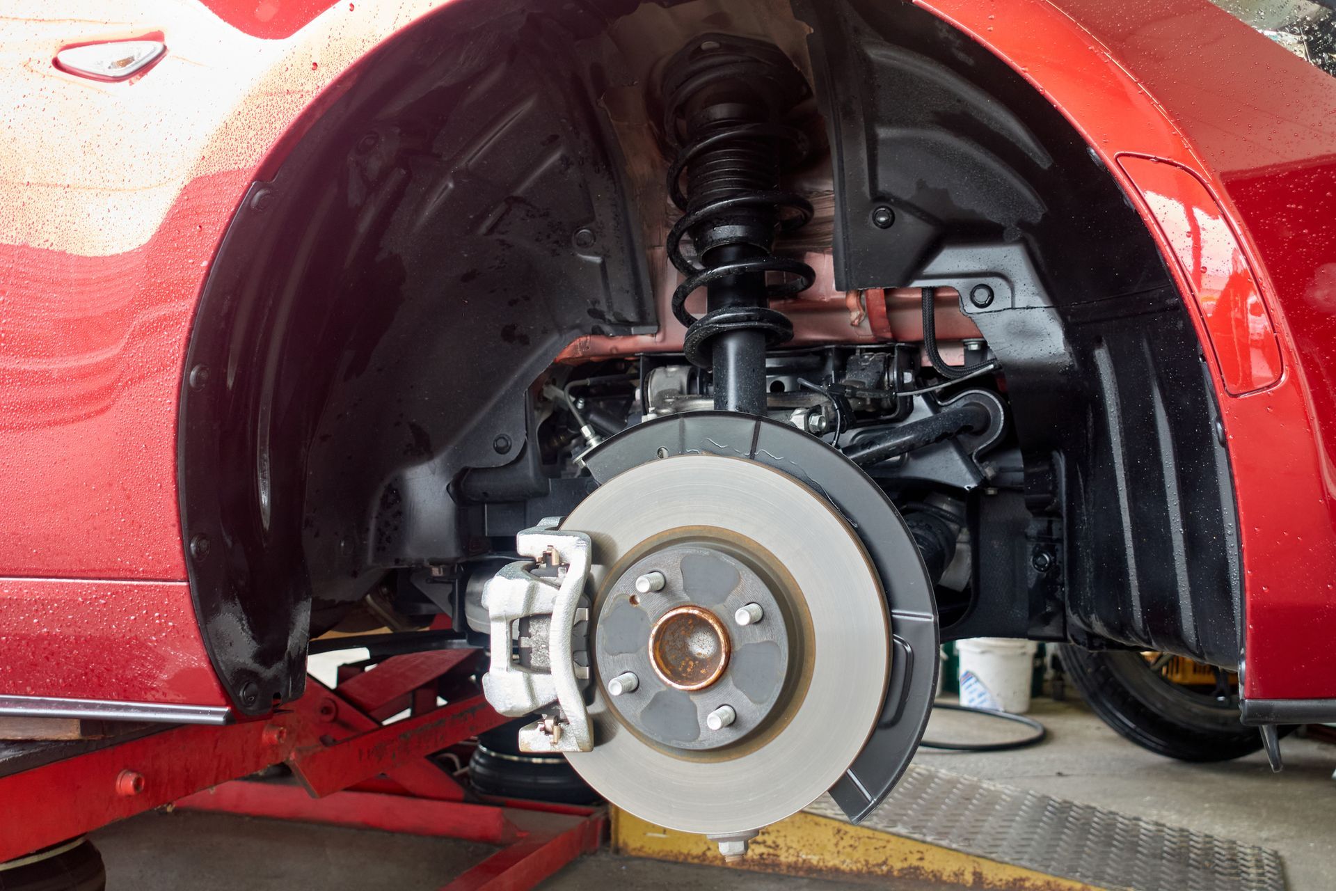 Red car's front wheel removed, revealing suspension, brake disc, and caliper. Car is on a jack, in a garage.