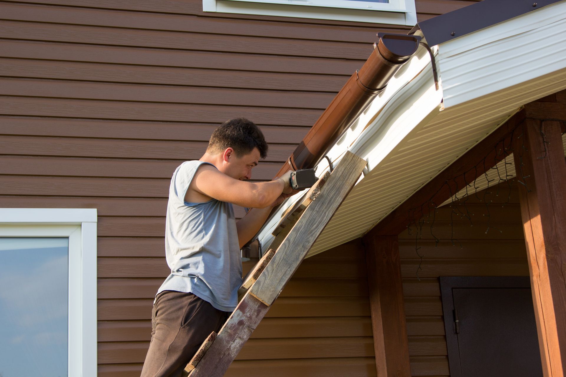 A man is standing on a ladder fixing a gutter on the side of a house.