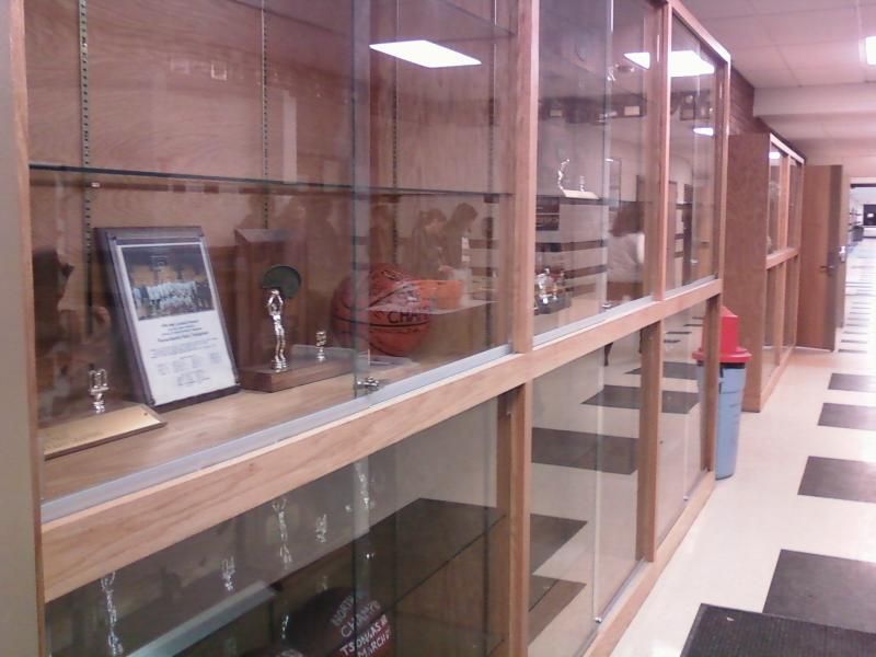 Glass display cases in a school hallway showcase trophies and awards. Wooden frames, cream walls, and a distant door.