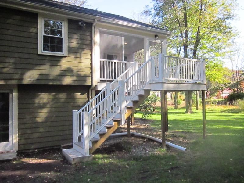 White deck with stairs attached to a green house. The deck is raised above the grassy lawn.
