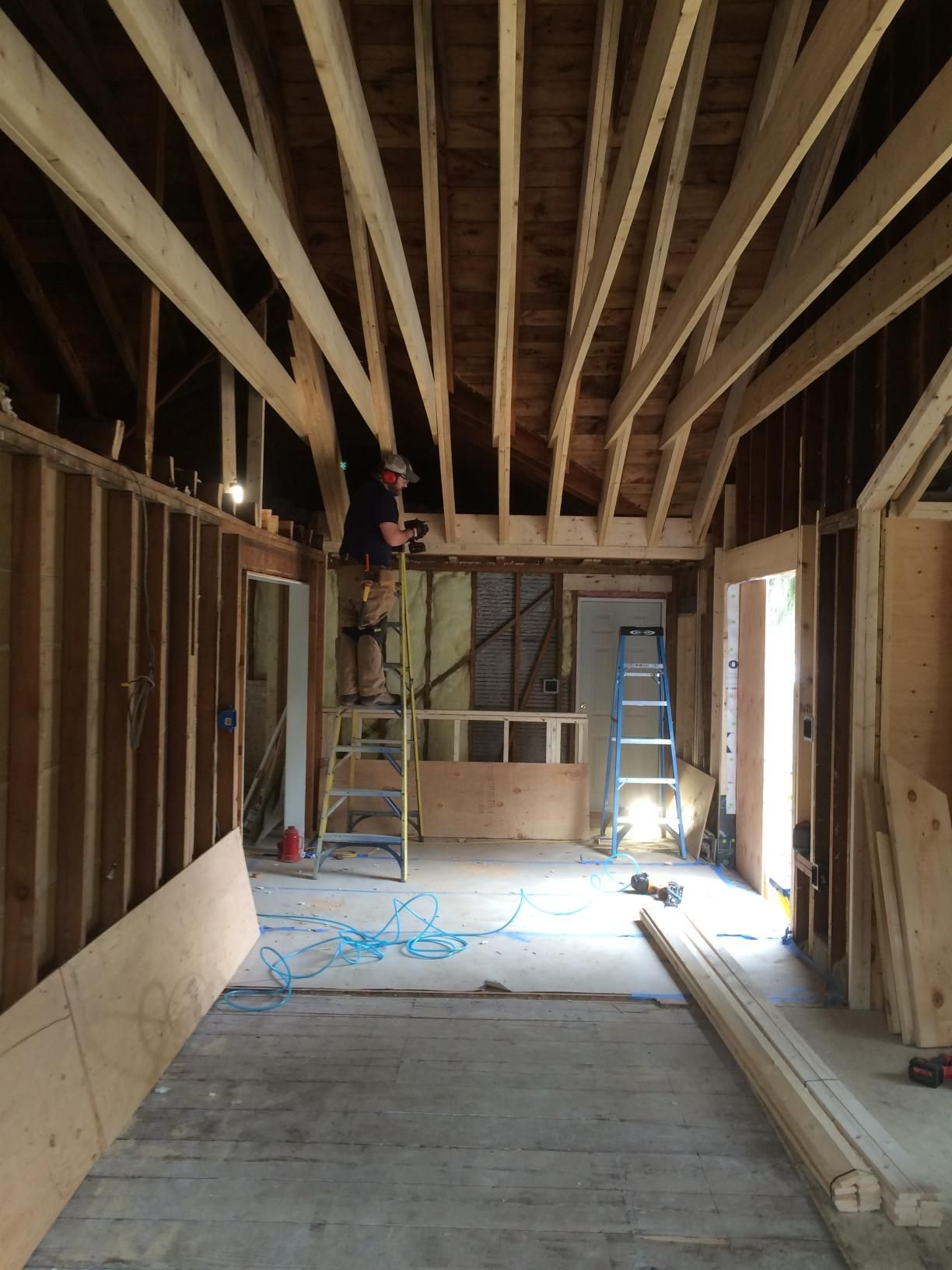 Interior of a room under construction with exposed wooden beams and studs, a person on a ladder.