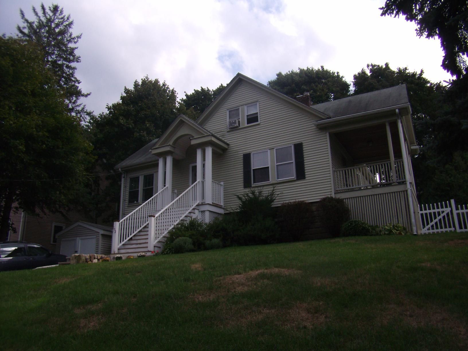 Two-story house with white siding, porch, and stairs, set on a grassy hill under an overcast sky.