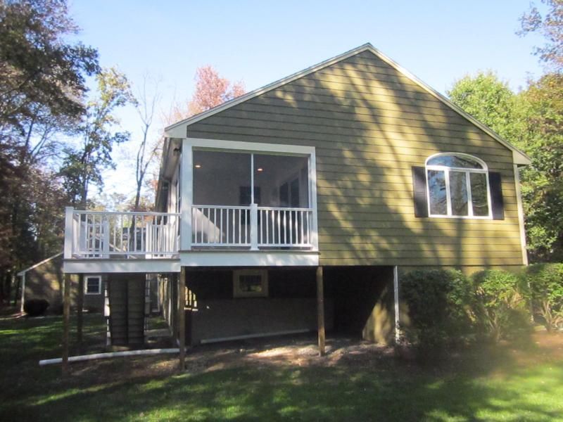 Green house with a deck, screened porch, white railings, and black shutters surrounded by trees.