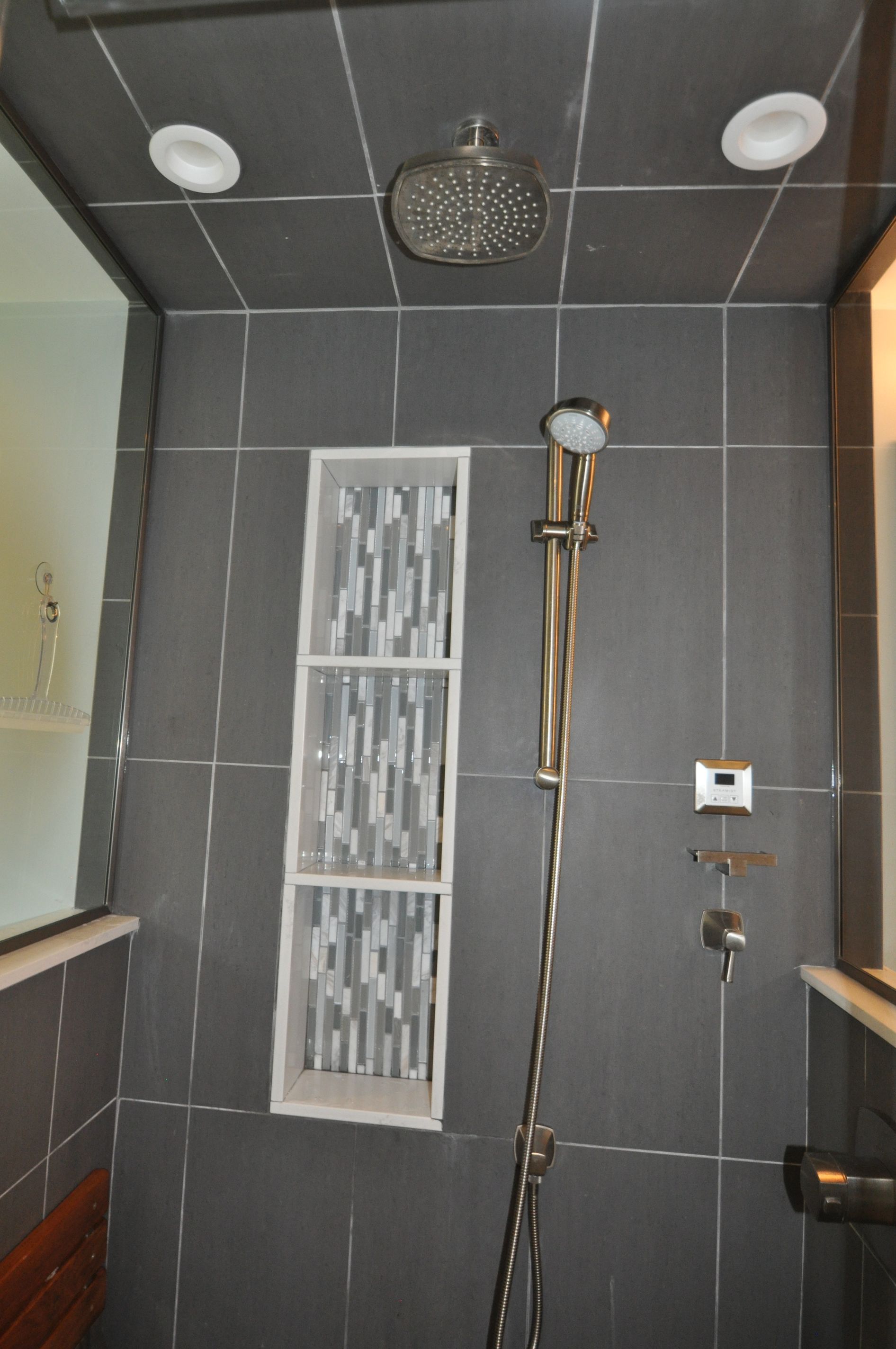 Gray tiled shower with rain showerhead, handheld shower, recessed shelf, and recessed lighting.