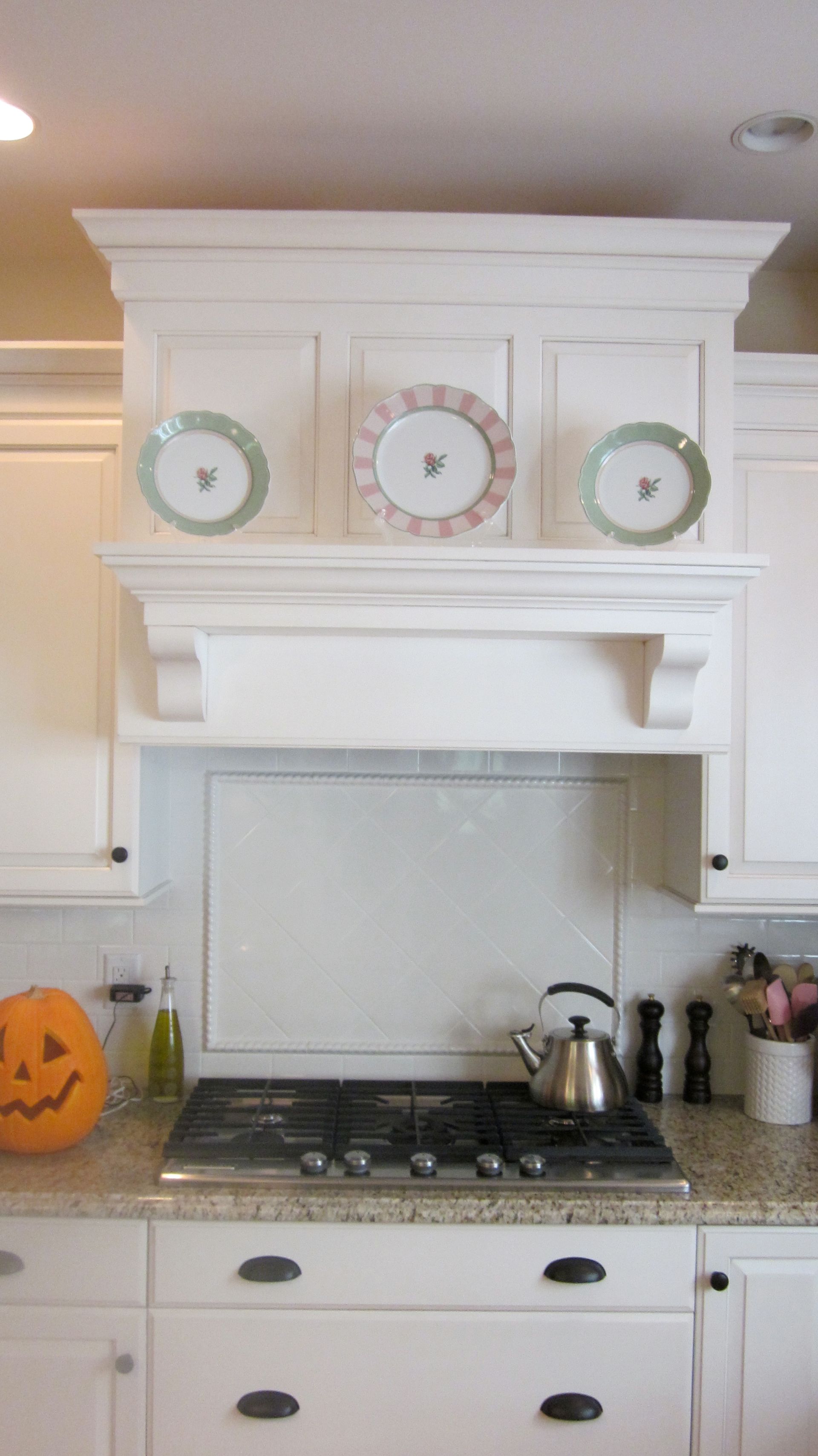 White kitchen range hood with decorative plates above a gas cooktop. A jack-o'-lantern is on the counter.