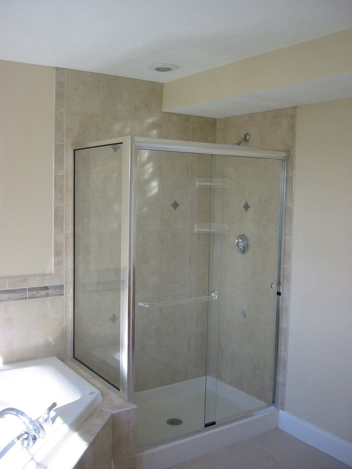 Corner shower with glass doors and beige tile, inside a bathroom with beige walls.
