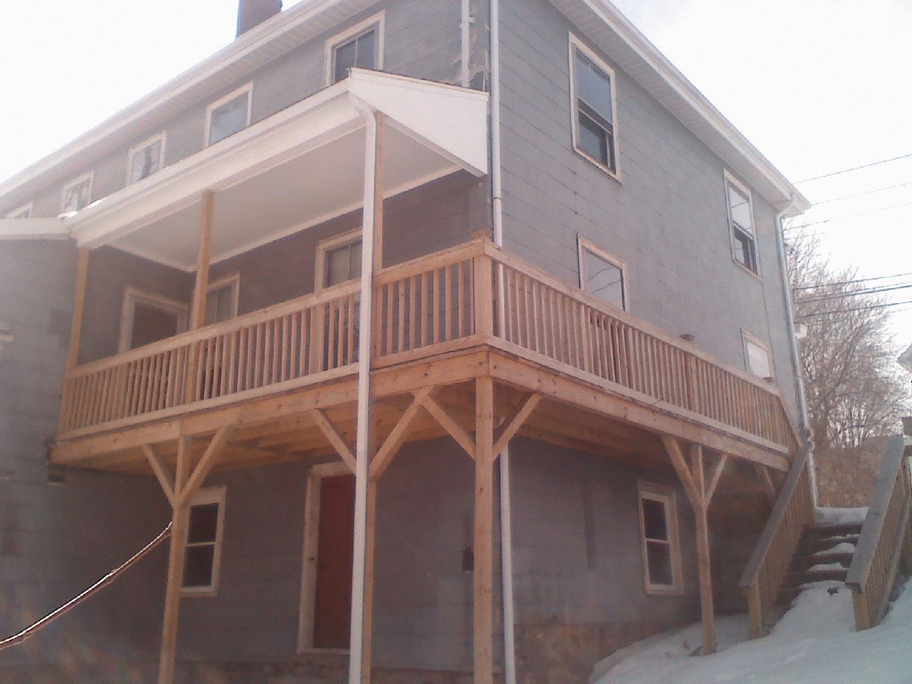 Two-story gray house with a wooden deck and stairs. Red door, windows, and a snowy ground are visible.