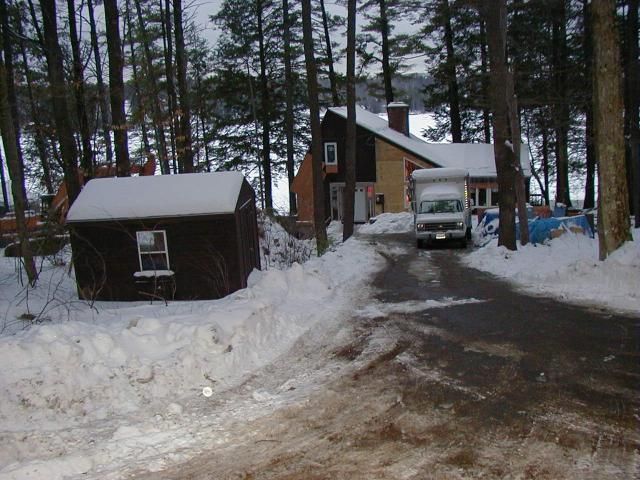 Winter scene: House with a shed on a snowy driveway; a van parked in front, trees surround the home.
