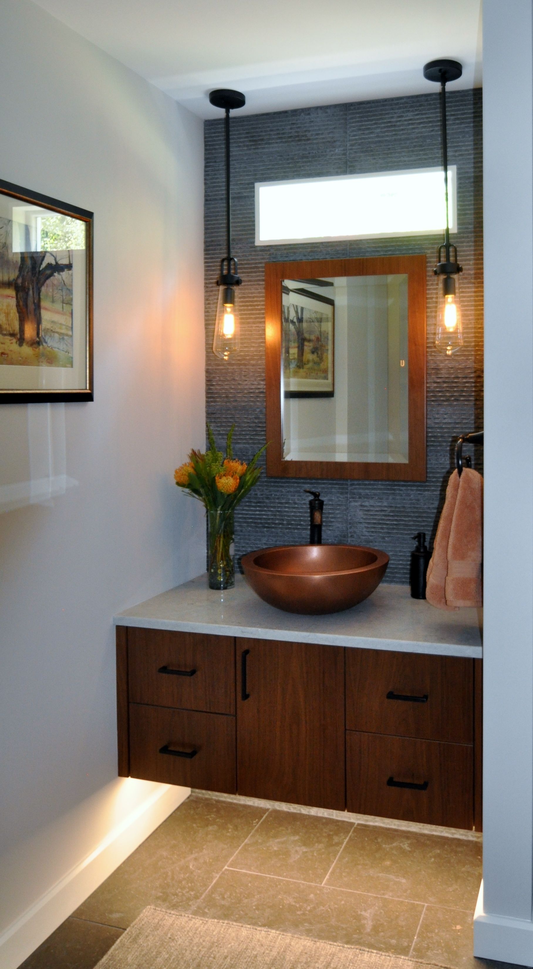Modern powder room with a copper sink, wood vanity, and pendant lights. Dark accent wall, stone floor.