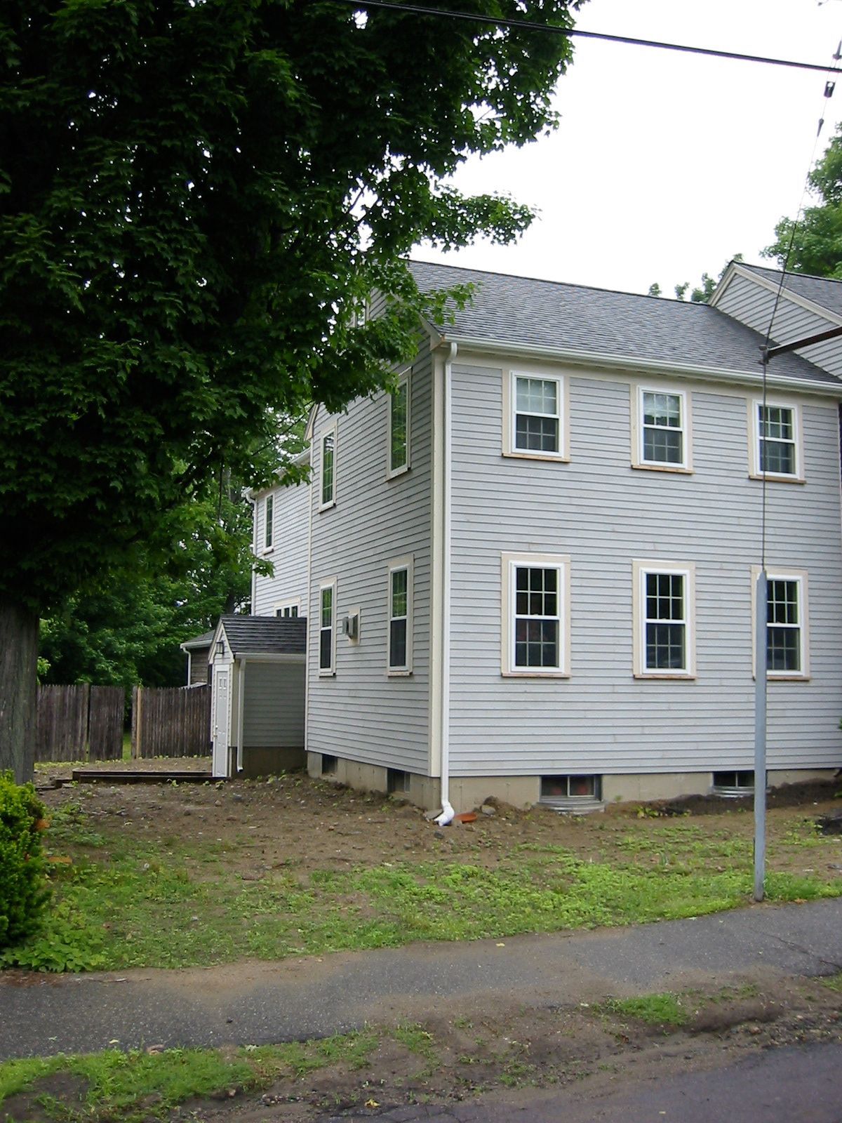 Two-story light gray house with white trim and multiple windows, green grass and trees, overcast sky.